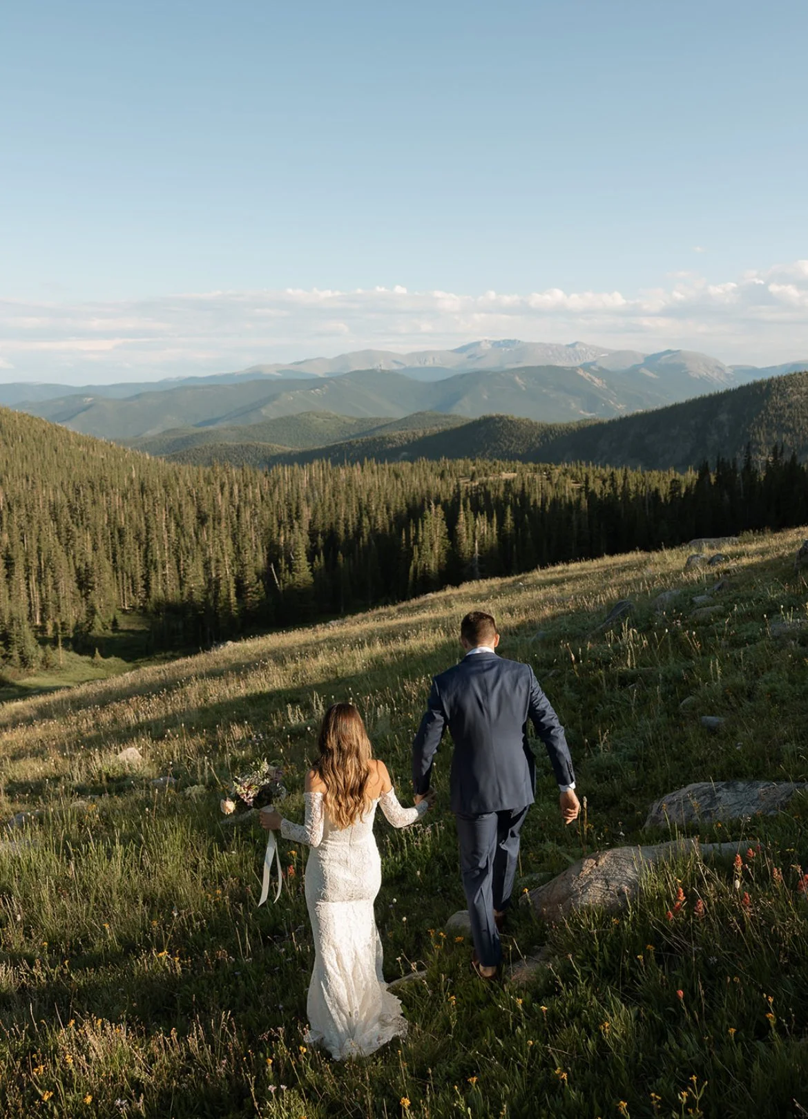 Couple in wedding attire walking through a scenic mountain landscape with a forest in the background.