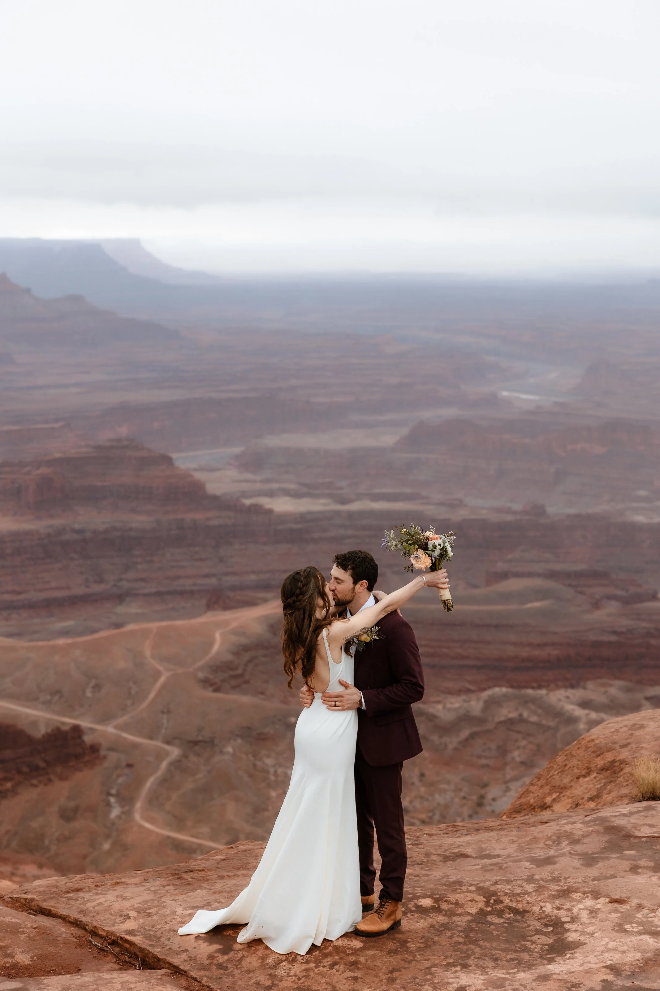 A couple kissing at a cliff's edge with a bouquet, overlooking a canyon landscape.