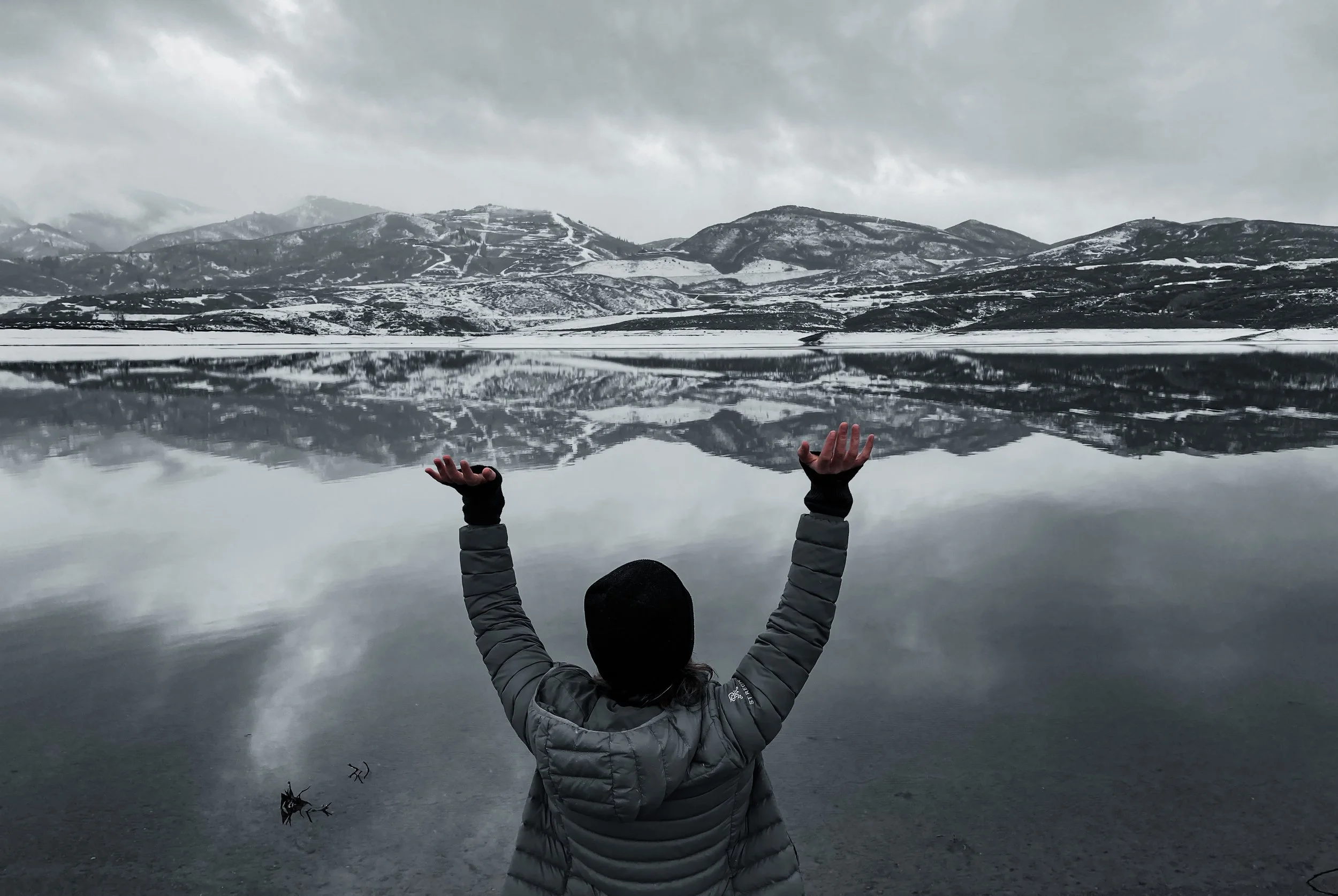 An artist wearing a gray jacket and black hat standing by a calm lake with arms raised, facing snow-covered mountains and cloudy sky, with the lake reflecting the landscape.