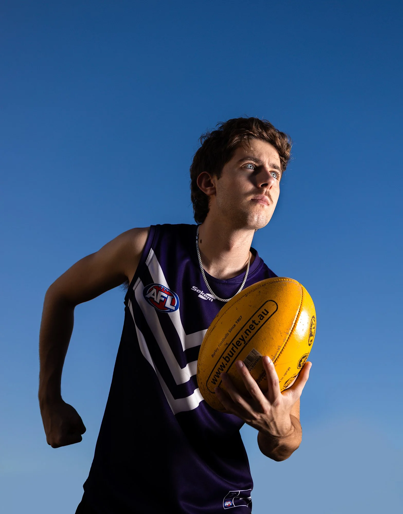 Young male Australian Rules football player holding a yellow football, wearing a purple jersey with AFL logo, standing against a clear blue sky.