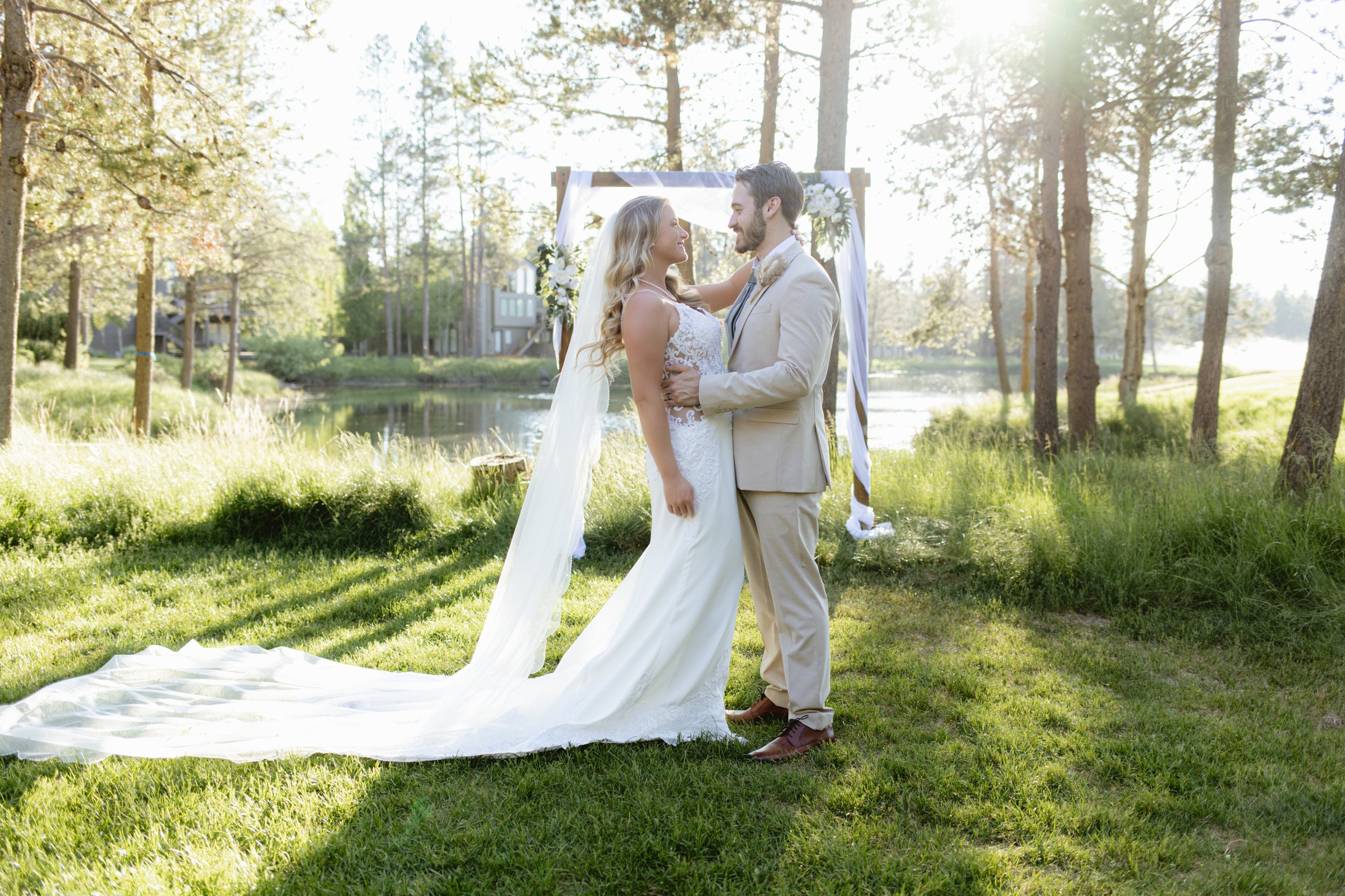 A bride and groom stand close together outdoors near a lake, facing each other on a sunny day. The bride wears a white lace wedding dress with a long train and veil, while the groom wears a light beige suit. There is a wooden wedding arch decorated w