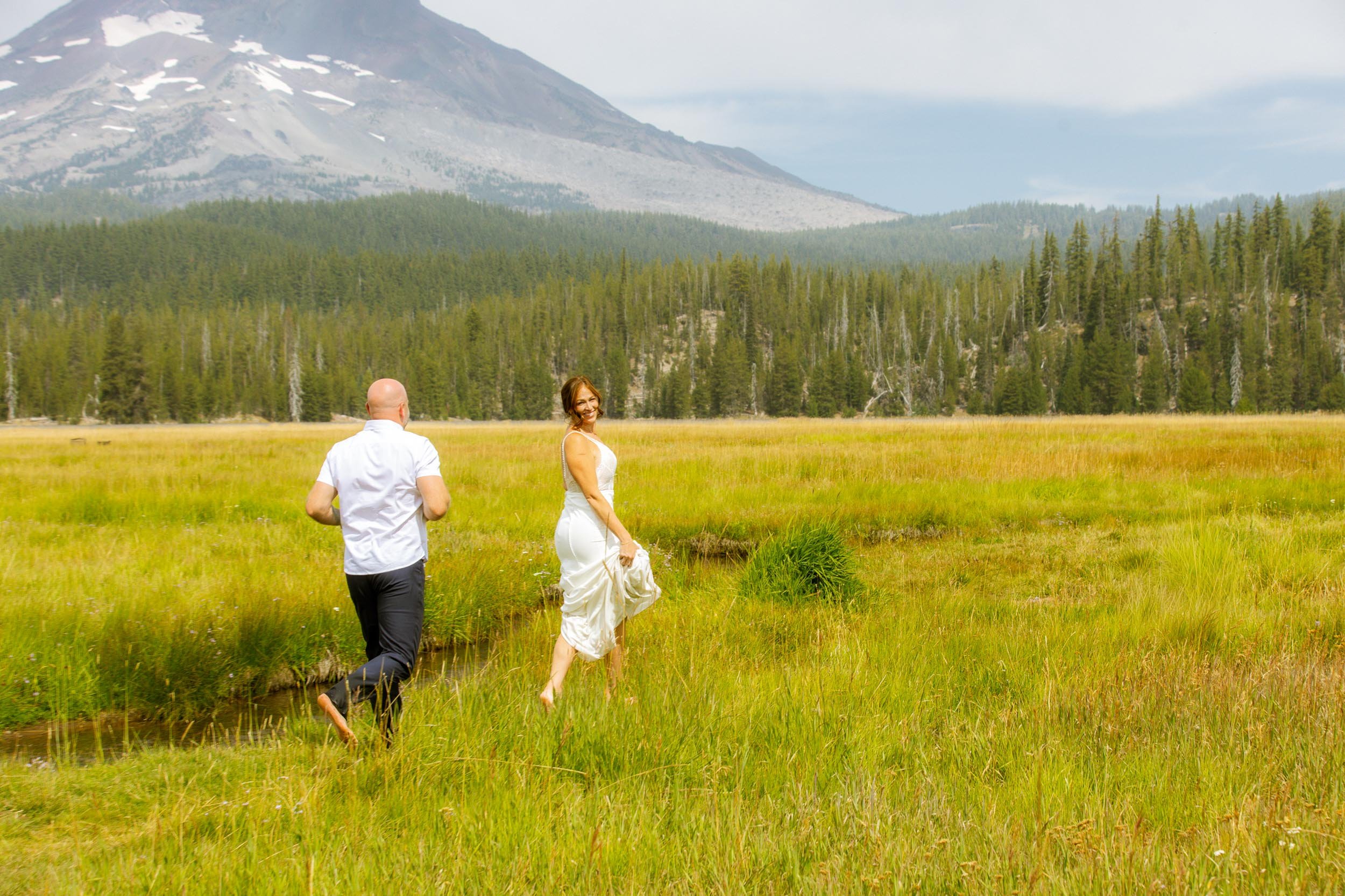 A bride and groom standing on a sandy shore by a calm lake with trees and mountains in the background during daytime.
