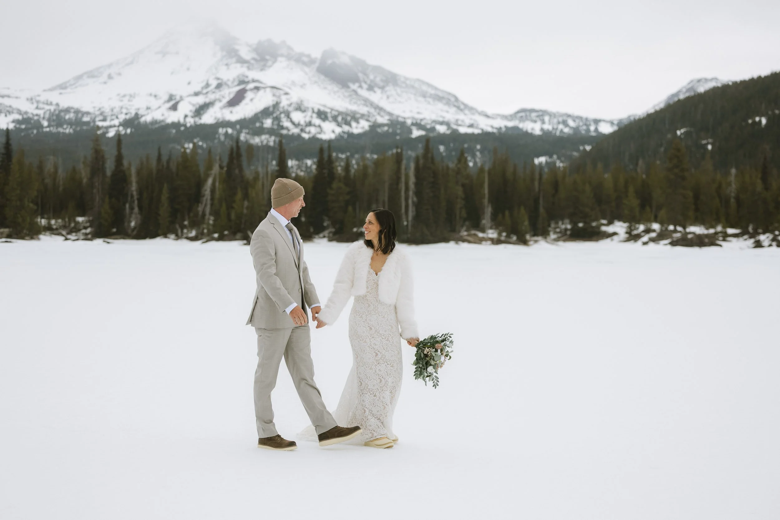 Couple in wedding attire holding hands in a snowy landscape with a forest and snow-capped mountains in the background.