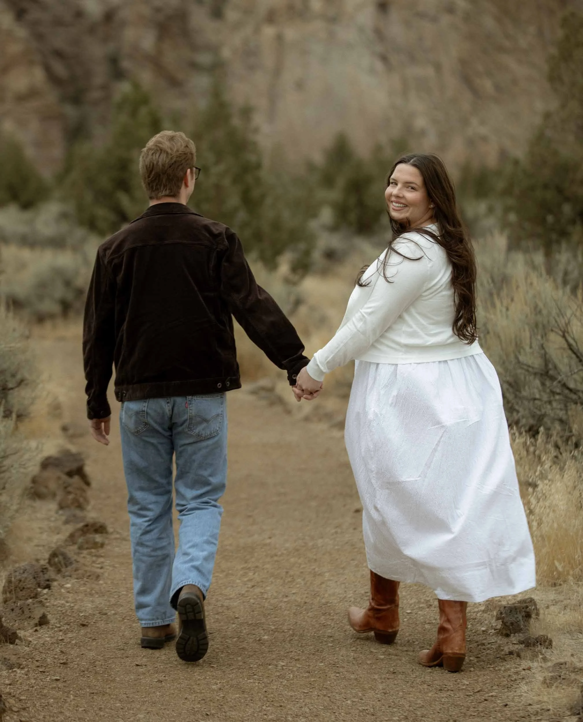A couple holding hands walking on a trail through a natural landscape with trees and mountains in the background.