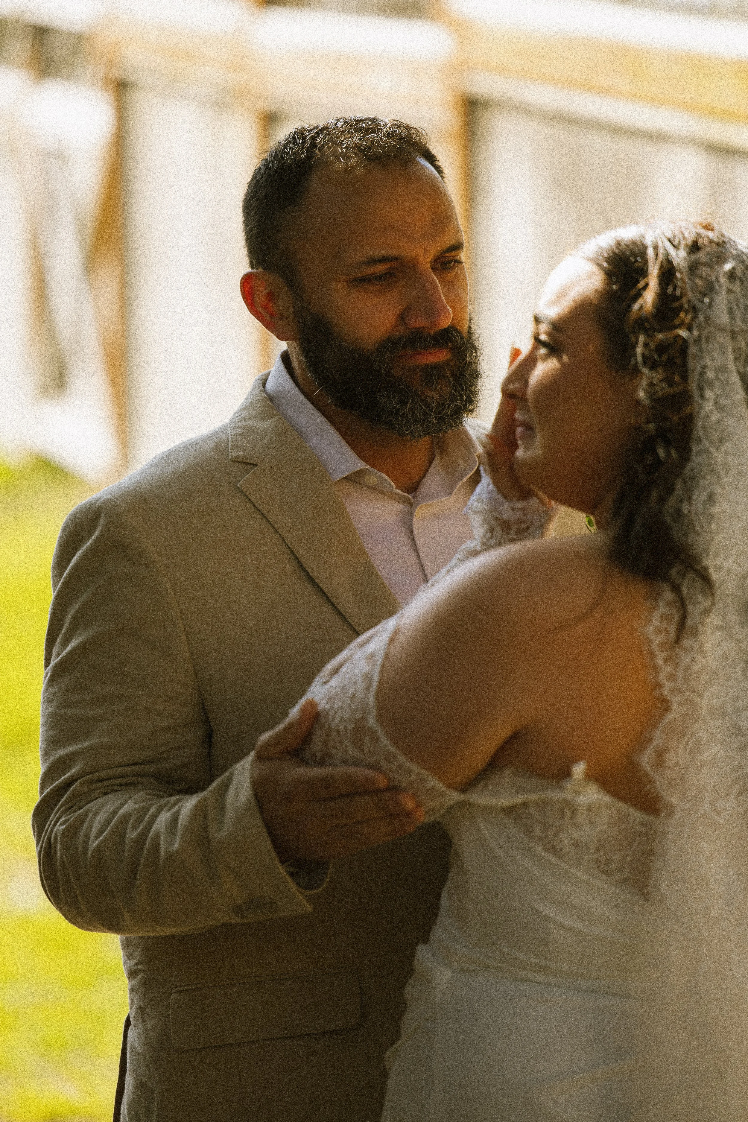 A man and woman are dancing closely at a wedding. The man has a beard and is dressed in a beige suit, while the woman is in a white lace wedding dress with a veil. They are looking into each other's eyes, sharing an intimate moment.