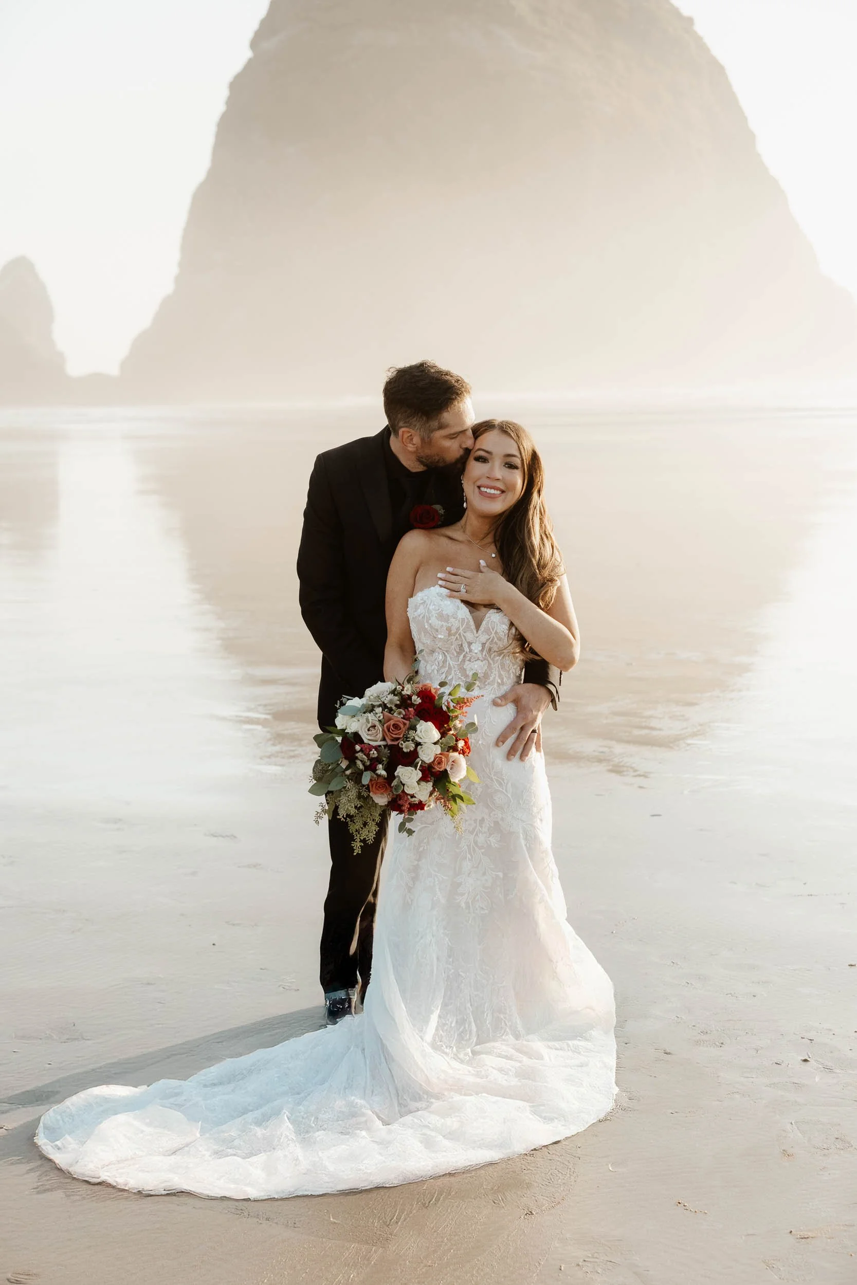 A bride and groom sharing a kiss by a lake with mountains in the background.