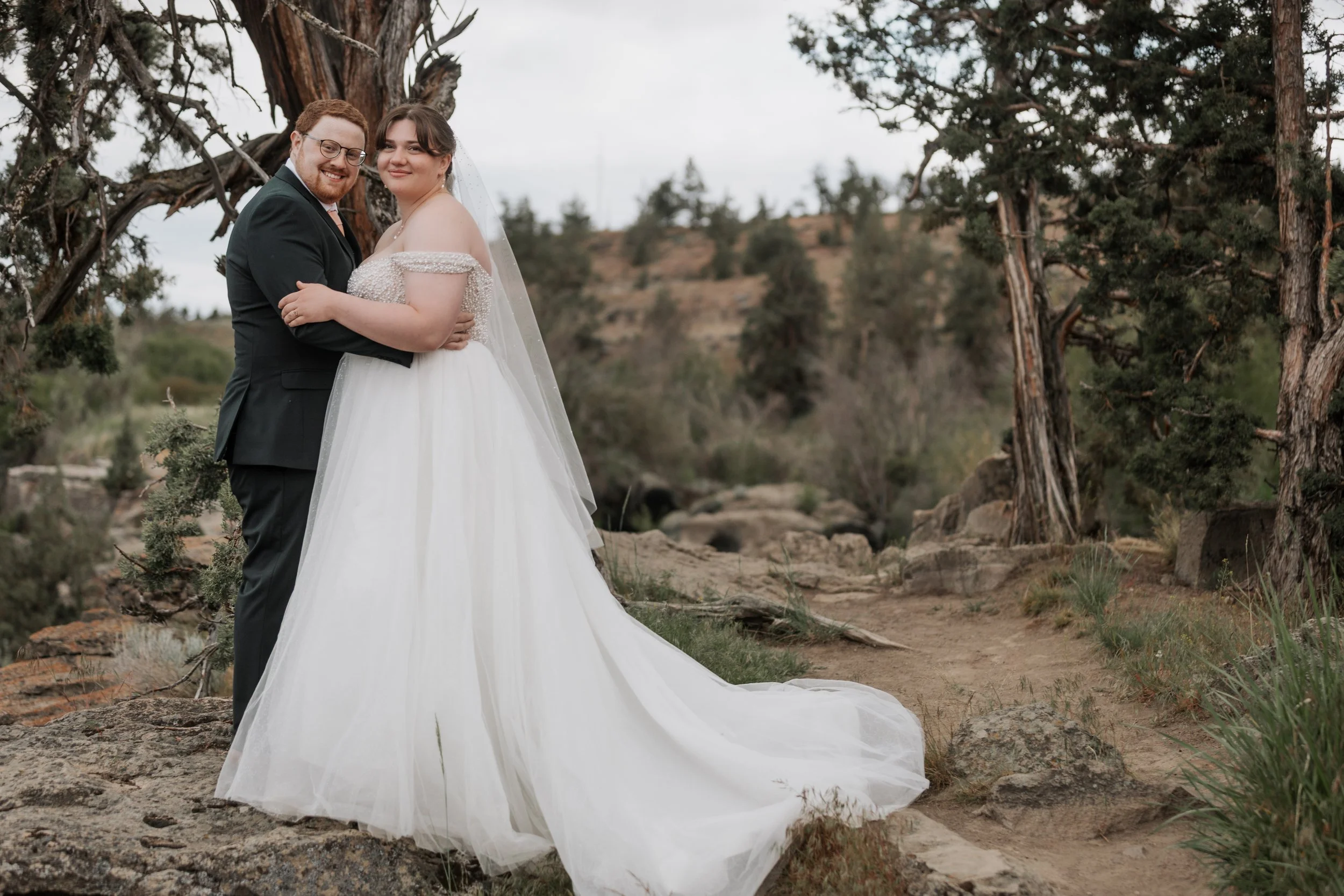 A couple dressed in wedding attire standing outdoors in a natural setting, hugging and smiling.