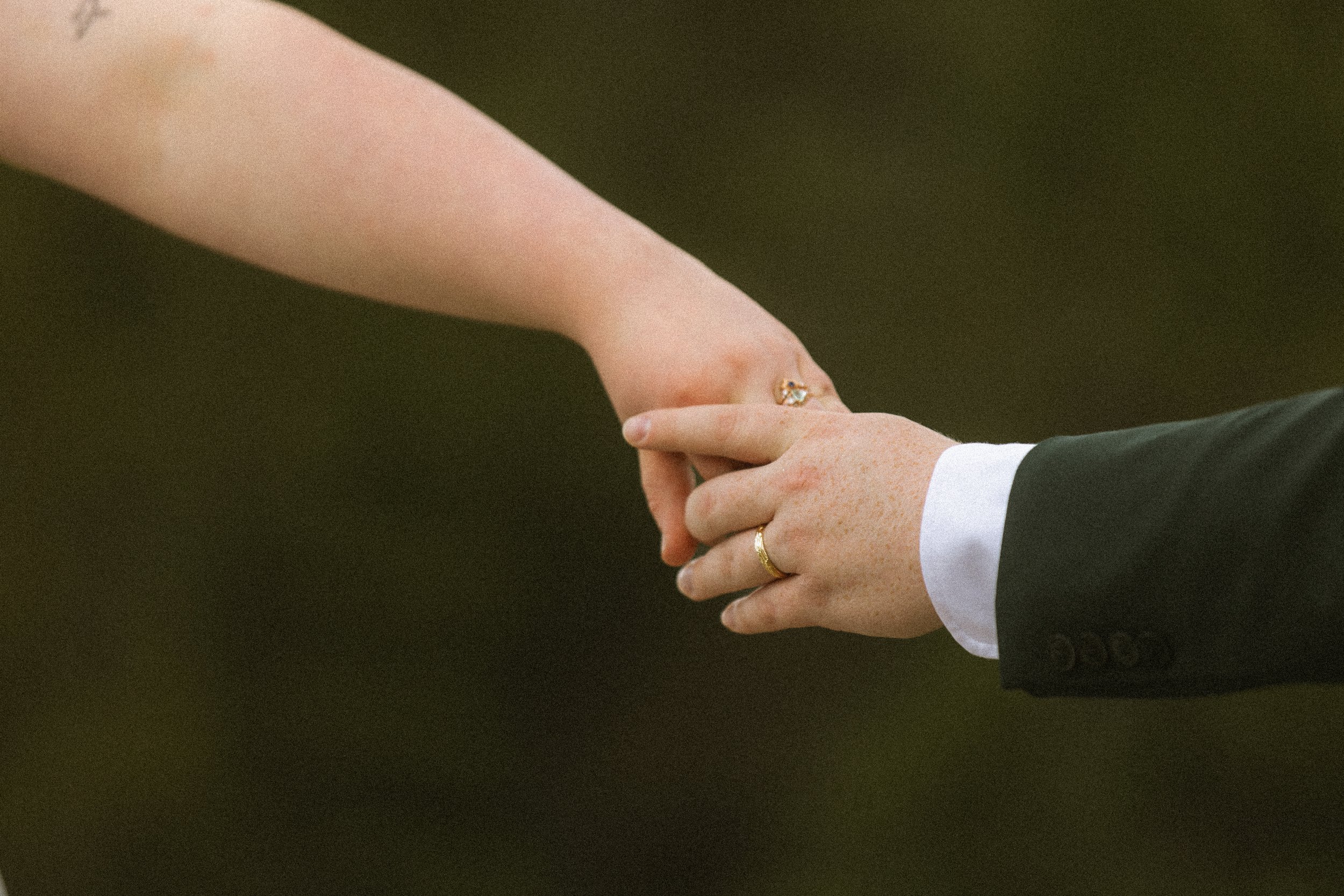 Close-up of a couple holding hands during their wedding ceremony, with the groom wearing a dark suit and the bride with a ring on her finger.