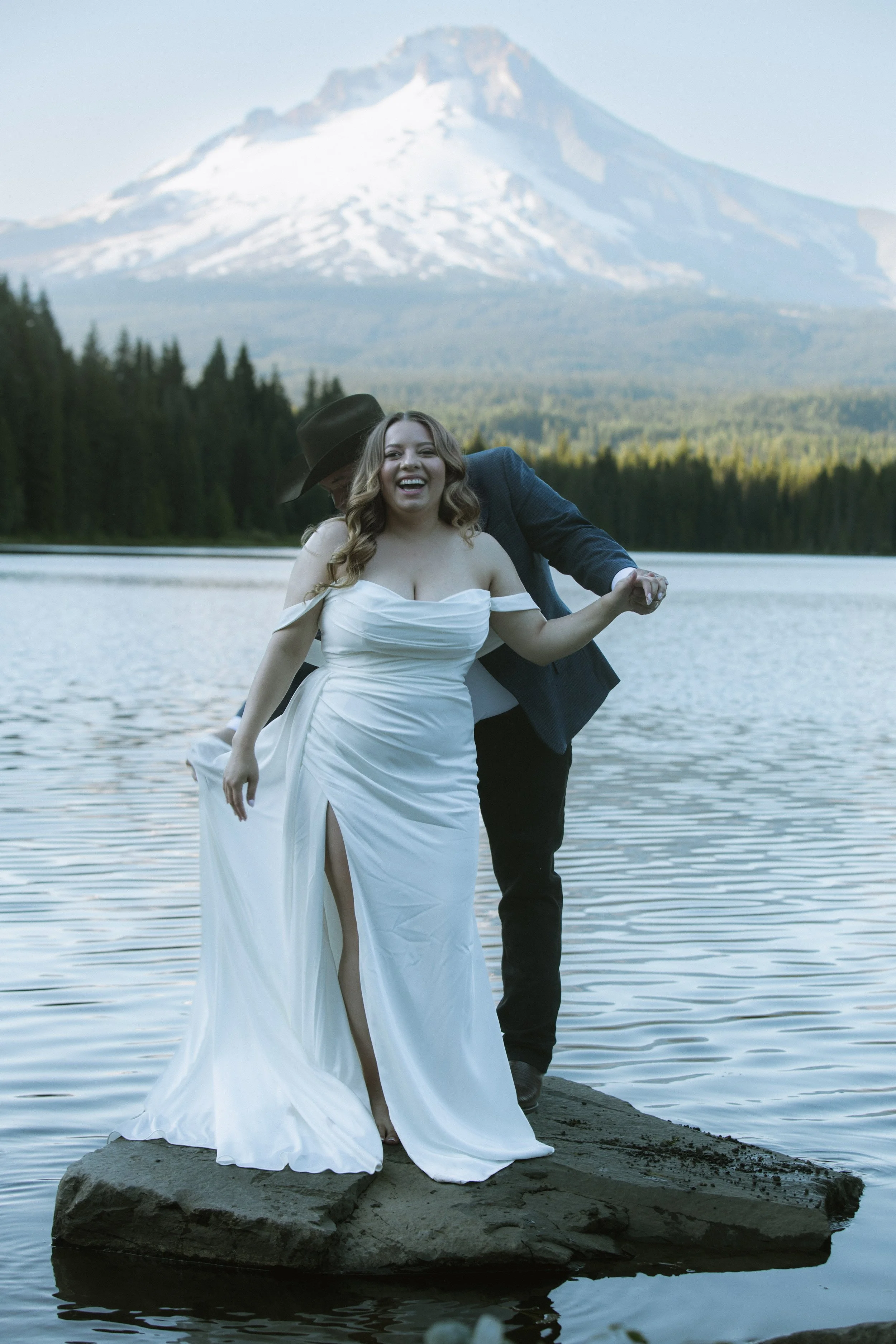 A couple dressed in wedding attire standing on a rock in a lake with a mountain in the background, smiling and holding hands.