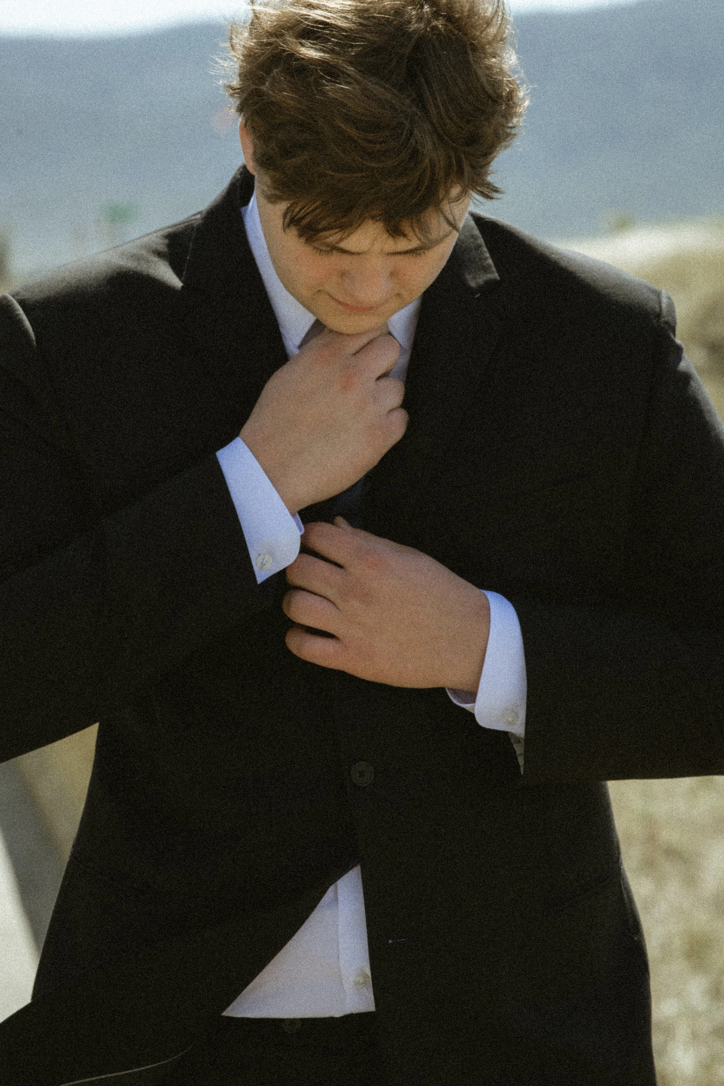 A person dressed in a dark suit with white shirt, adjusting the collar or tie, outdoors with a beach and ocean in the background.