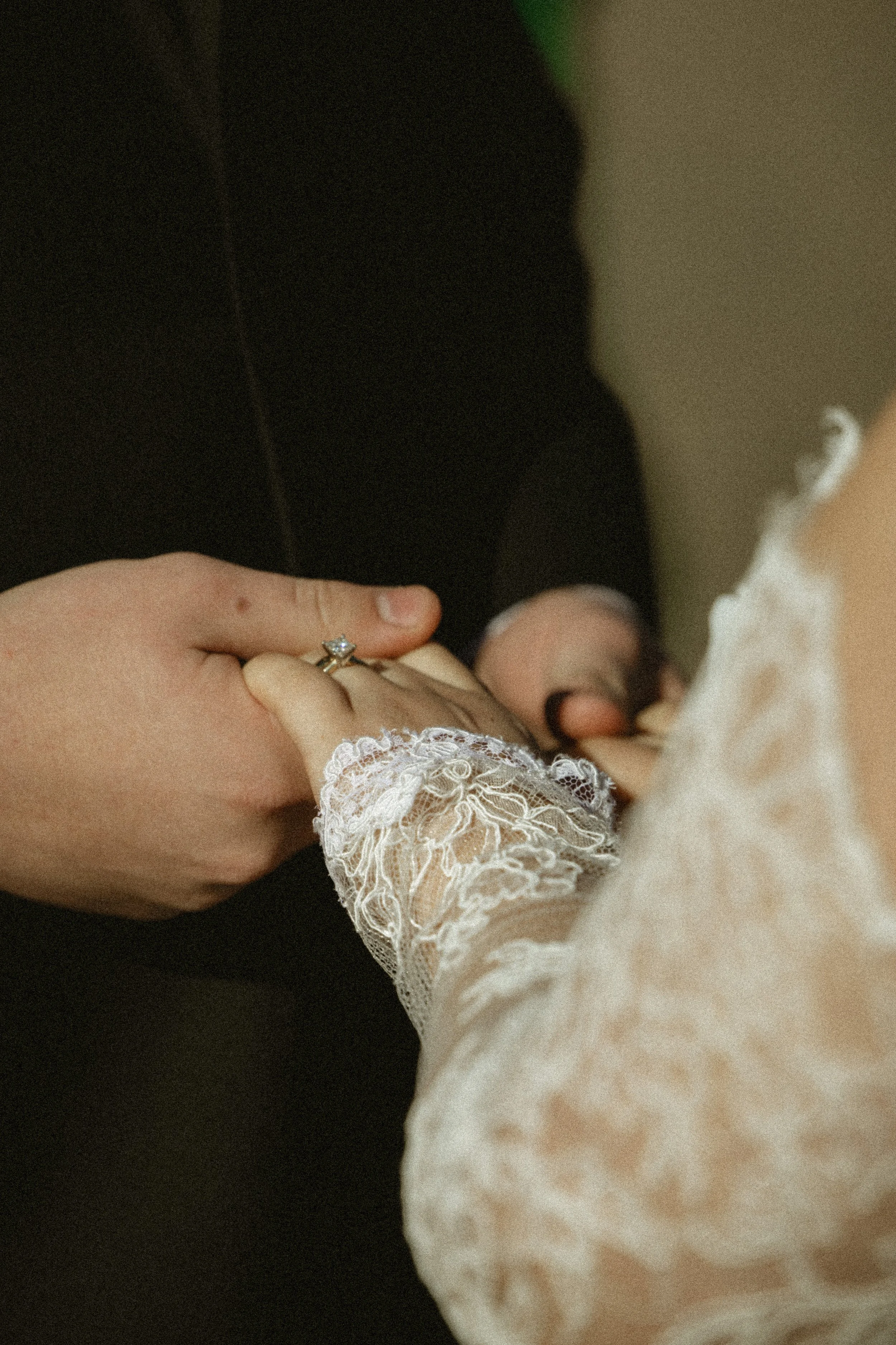 A person in a dark suit holding a baby's hand, which is dressed in a lace sleeve of a cream-colored garment.