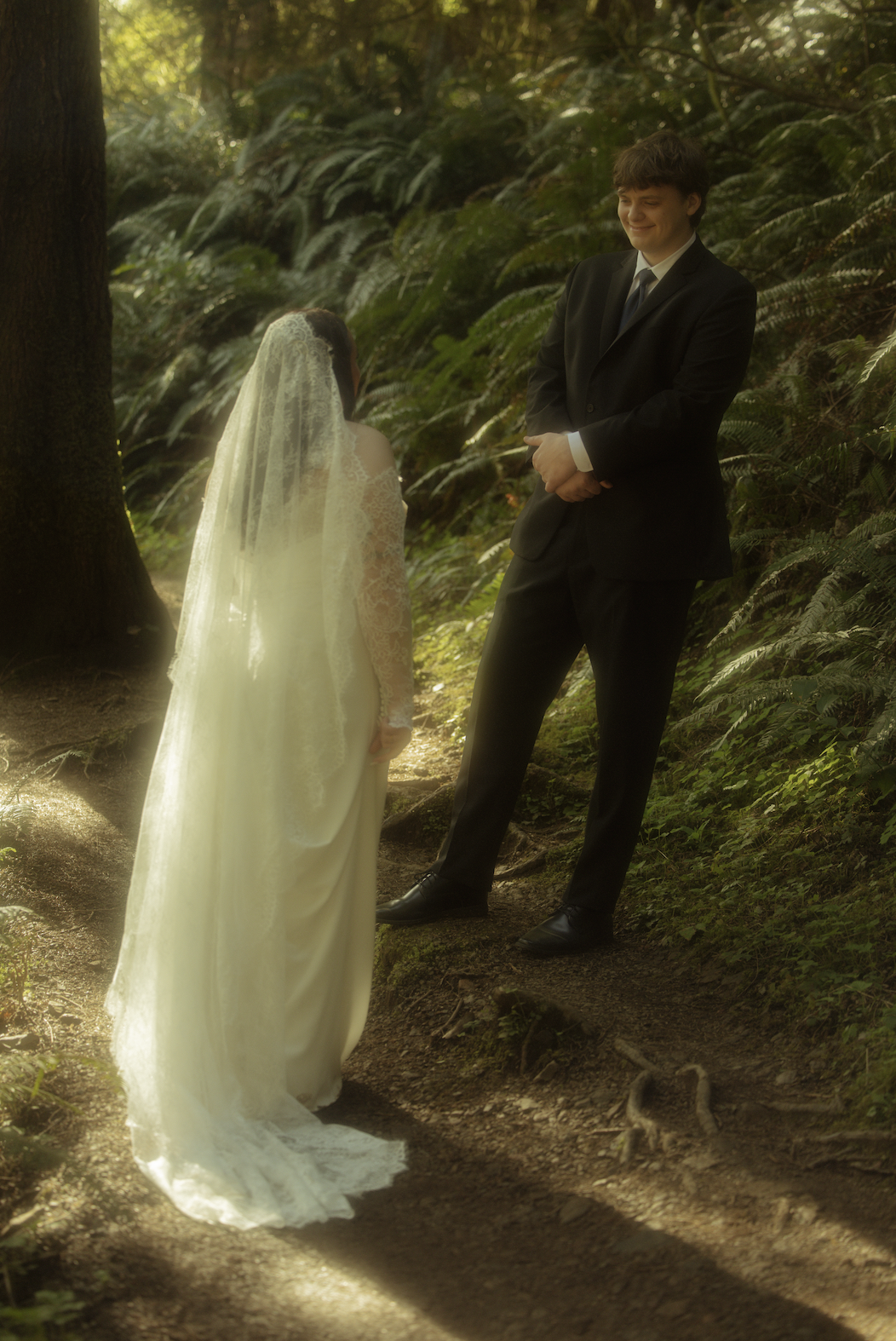 A bride and groom standing on a forest trail during daytime, with the groom dressed in a black suit and the bride in a white wedding gown with lace sleeves and a veil, facing each other amidst lush greenery.