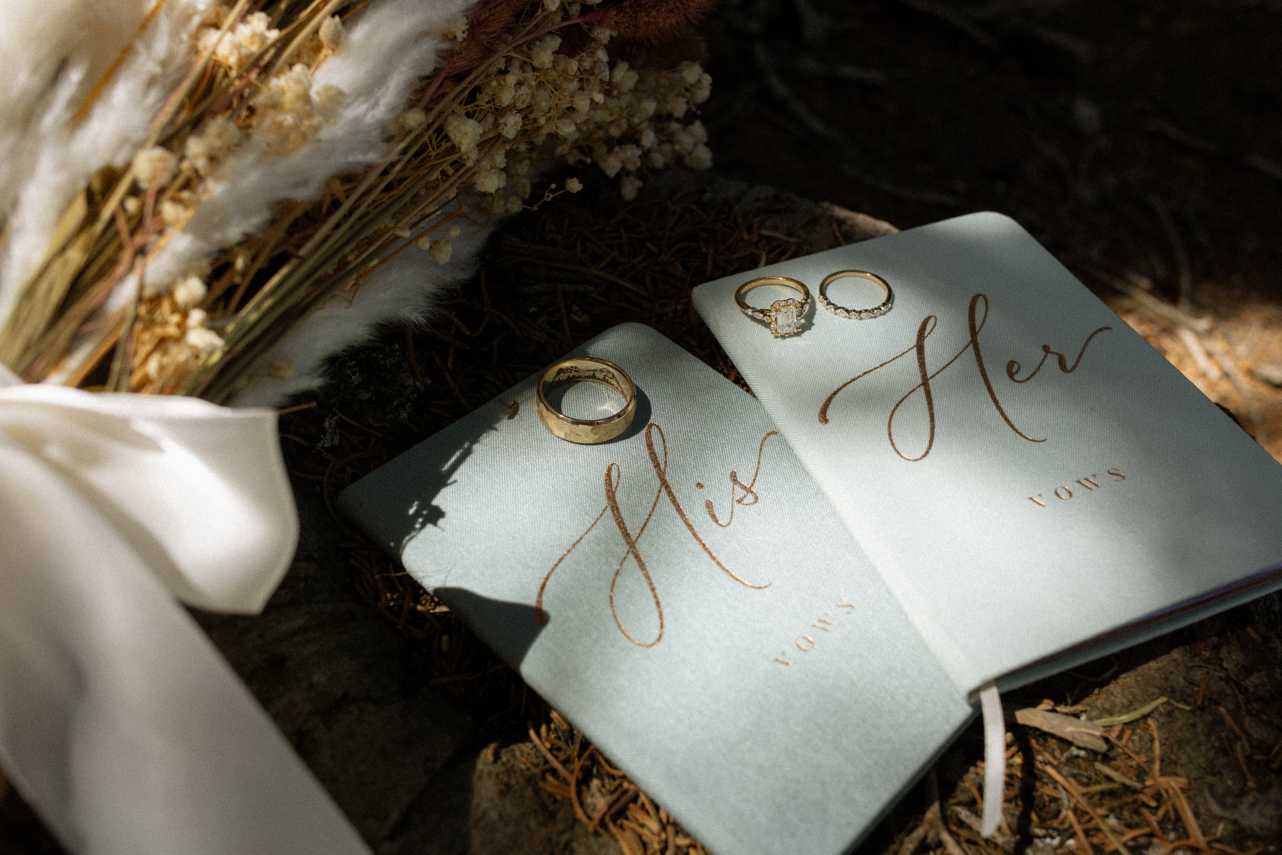 Two open wedding vow books with gold lettering: one labeled 'His' and the other 'Her,' with wedding rings placed on top, surrounded by dried flowers and a white ribbon.