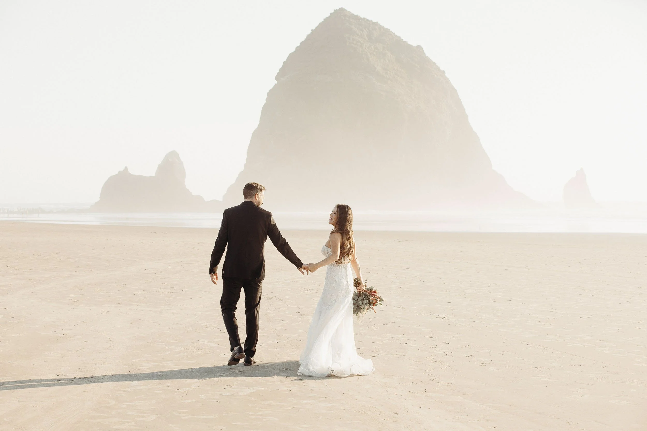 A bride and groom sharing a tender moment outdoors during their wedding, with the bride gazing up at the groom who is smiling softly. The groom is dressed in a light gray suit with a white tie and boutonniere, and the bride is holding a bouquet of colorful flowers.