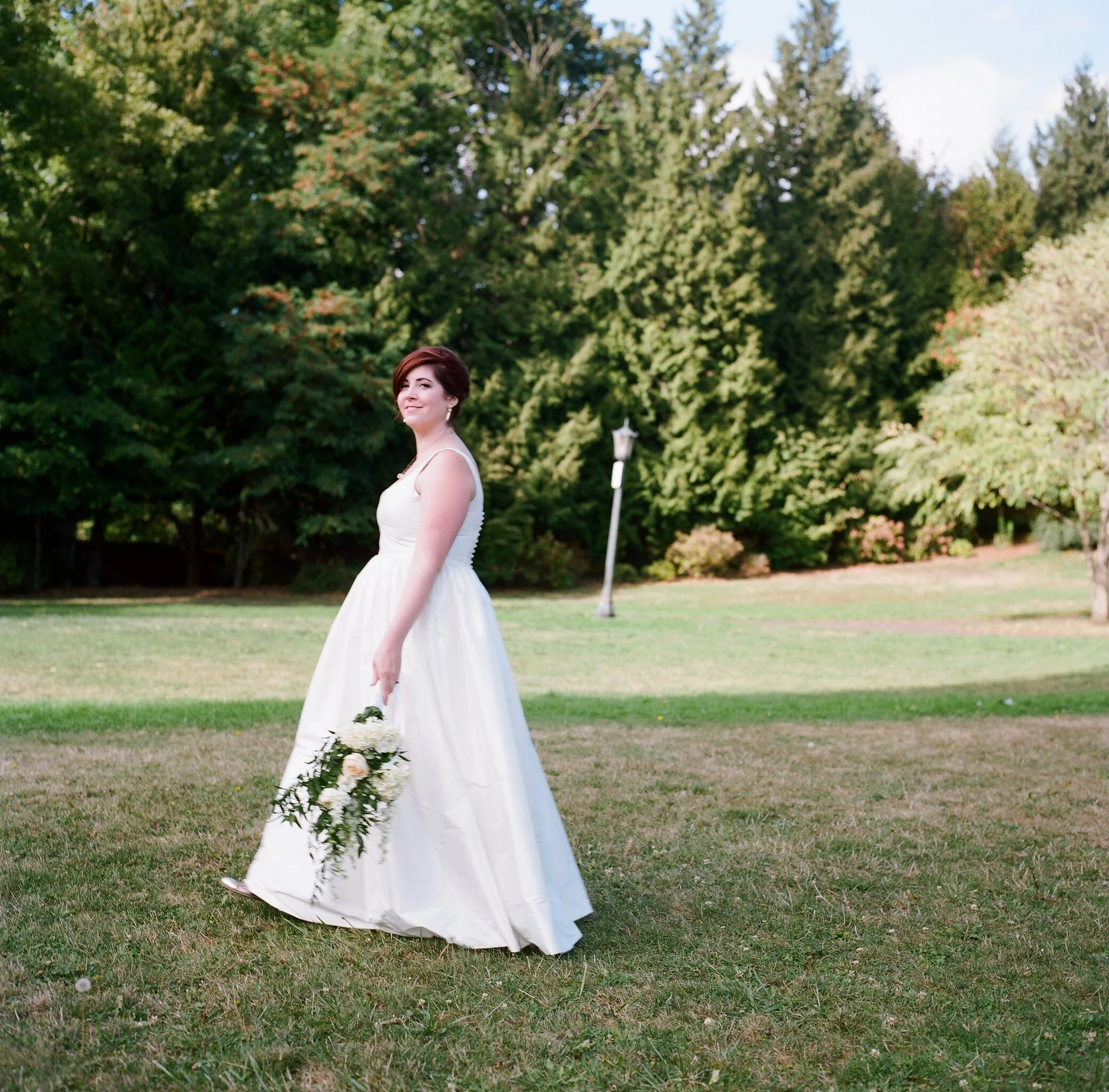 A woman in a white wedding dress holding a bouquet of flowers stands on a grassy field with trees in the background.