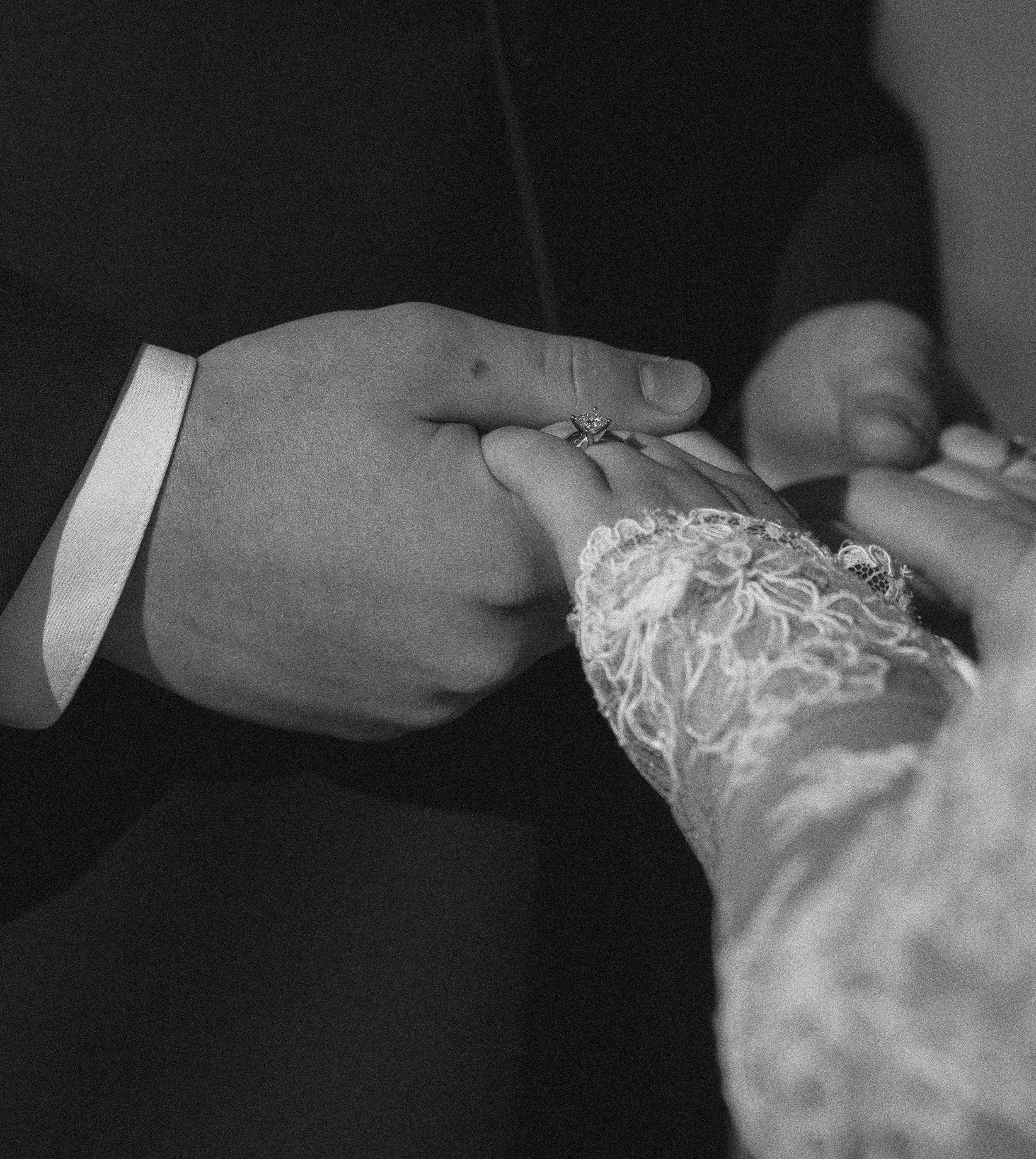 Close-up of hand with a ring on the finger, wearing lace clothing, against a dark background.