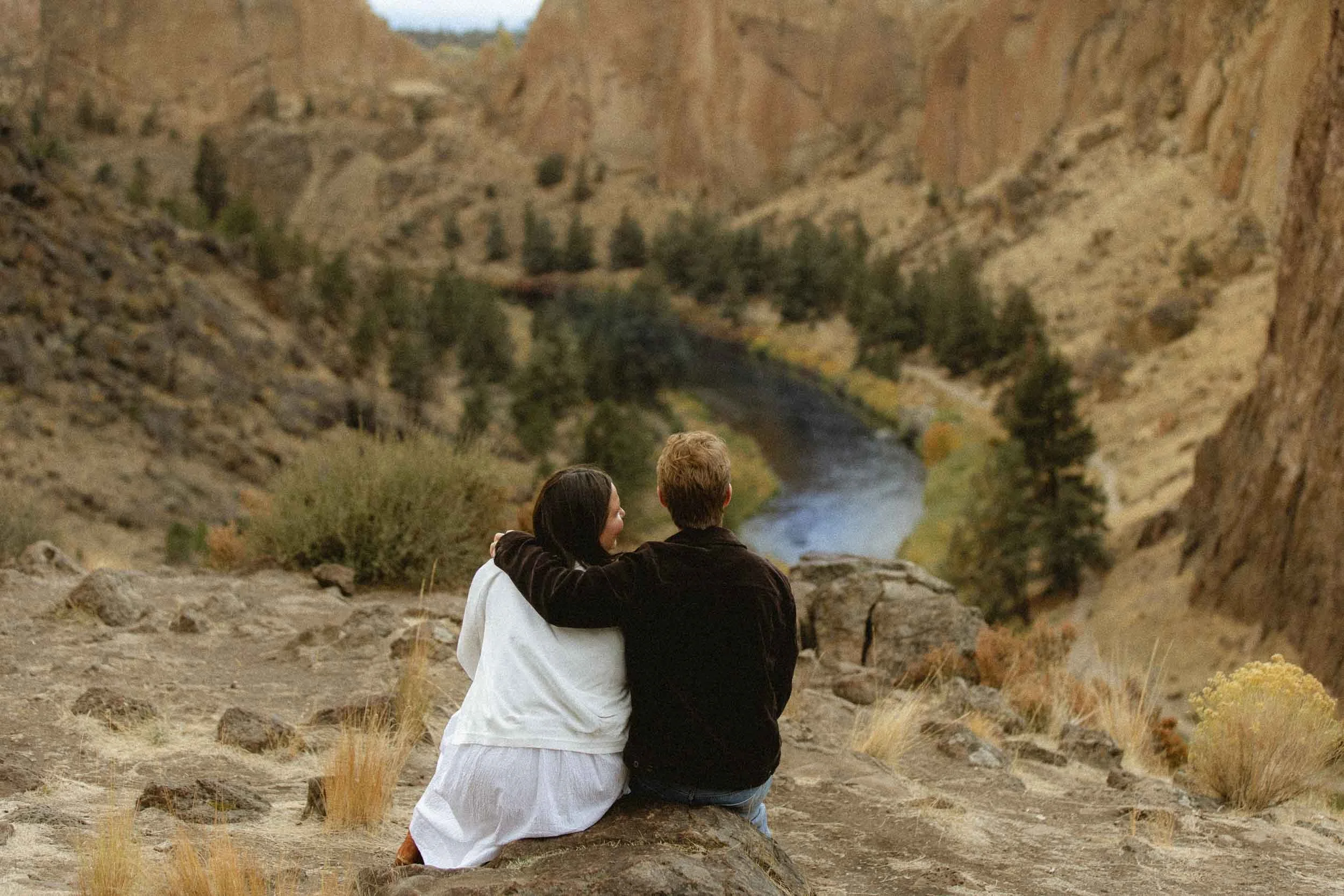 A couple sitting on a rock, embracing and looking at a river and canyon landscape.