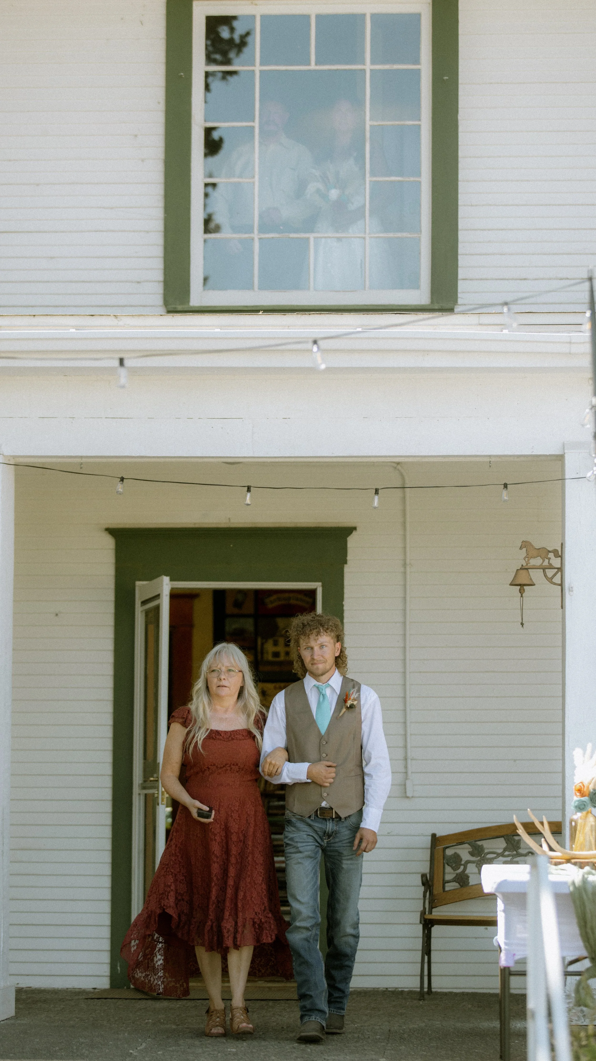 A young man with curly hair and a woman with long gray hair walk out of a house. The woman wears a red lace dress and glasses, while the man wears a vest, white shirt, and tie, with a boutonniere. The house has white siding, a green trim around the d