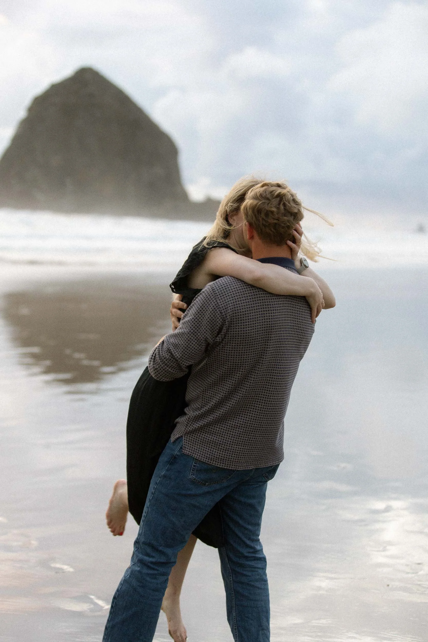 A couple embracing on a beach with a large rock formation in the background.