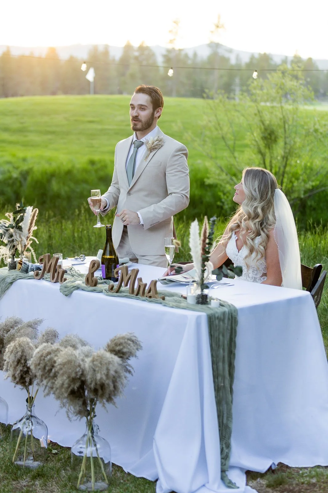 A man in a beige suit giving a speech at a wedding reception outdoors, with a bride sitting at the table beside him. The table is decorated with greenery, pampas grass, and a wooden sign reading 'Mr & Mrs'. There are glasses, bottles, and floral arrangements on the table, and the backdrop features lush green fields and trees with the setting sun.