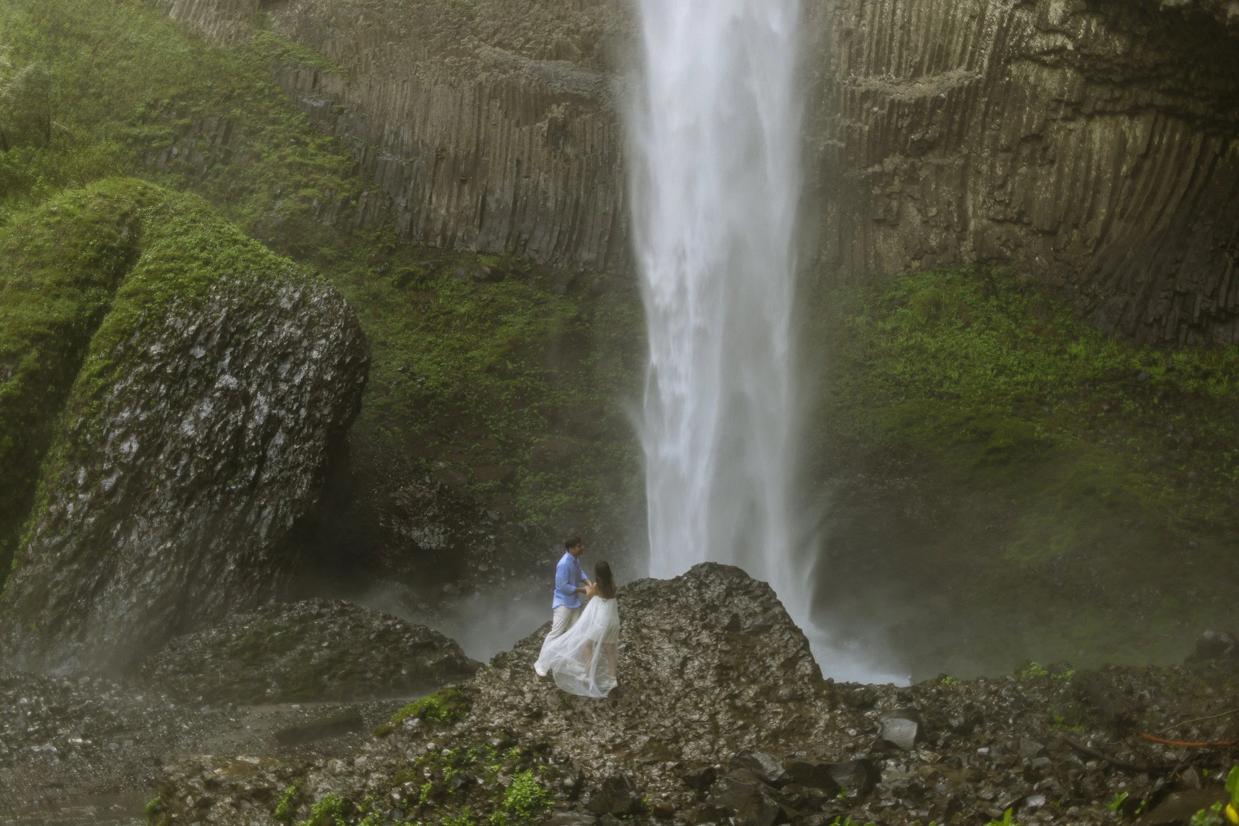 A couple standing under a waterfall in a lush green setting, similar to a scene from a movie or photo shoot.