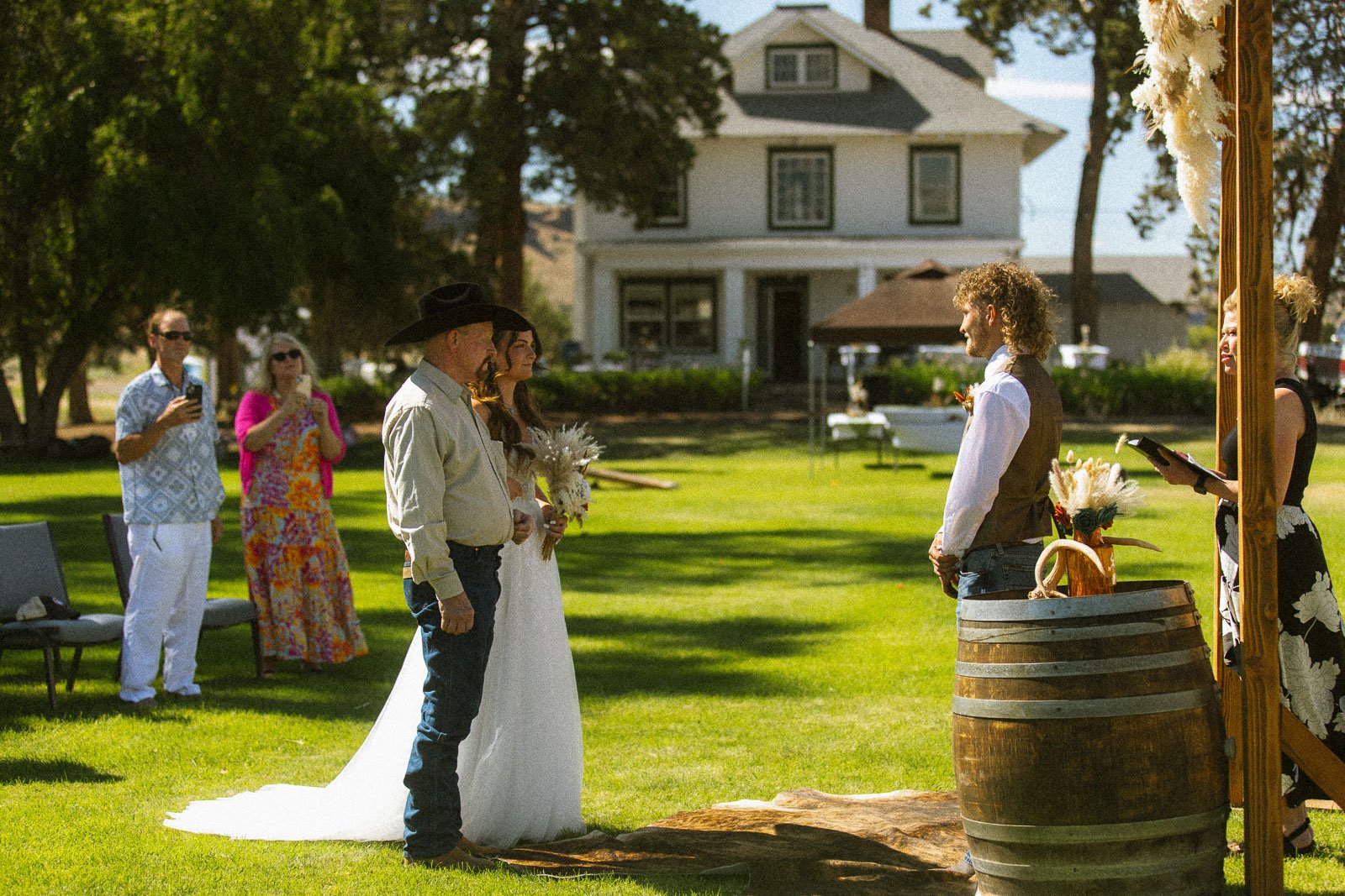A wedding ceremony taking place outdoors on a sunny day with the bride and groom standing in front of a minister. The bride is wearing a white gown and holding a bouquet, while the groom is in a cowboy hat, shirt, and jeans. Guests are standing and watching, some taking photos, with large trees and a house in the background.