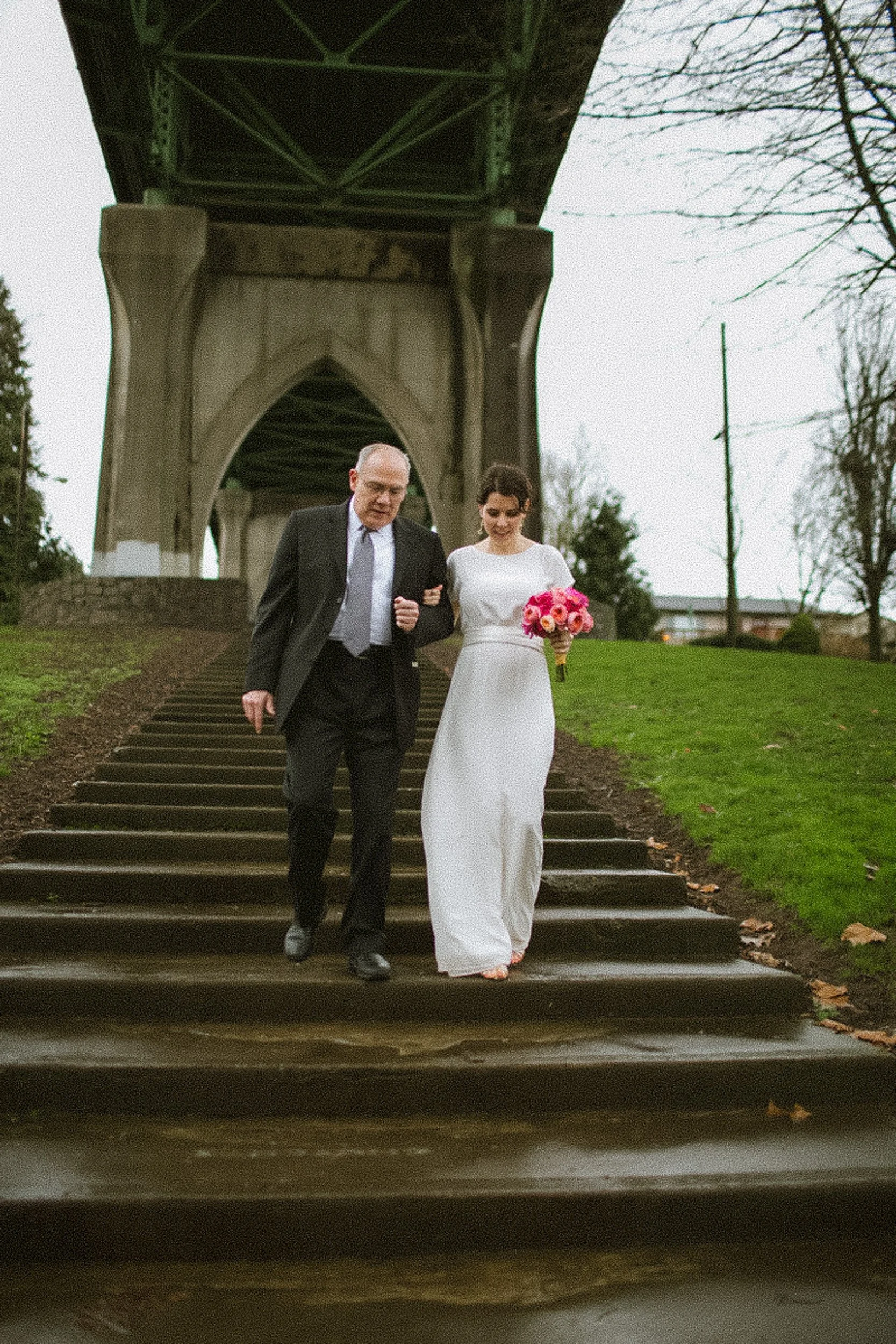 A bride in a white wedding dress holding a pink bouquet walks down outdoor stairs with an older man in a suit, under a bridge, on a cloudy day.