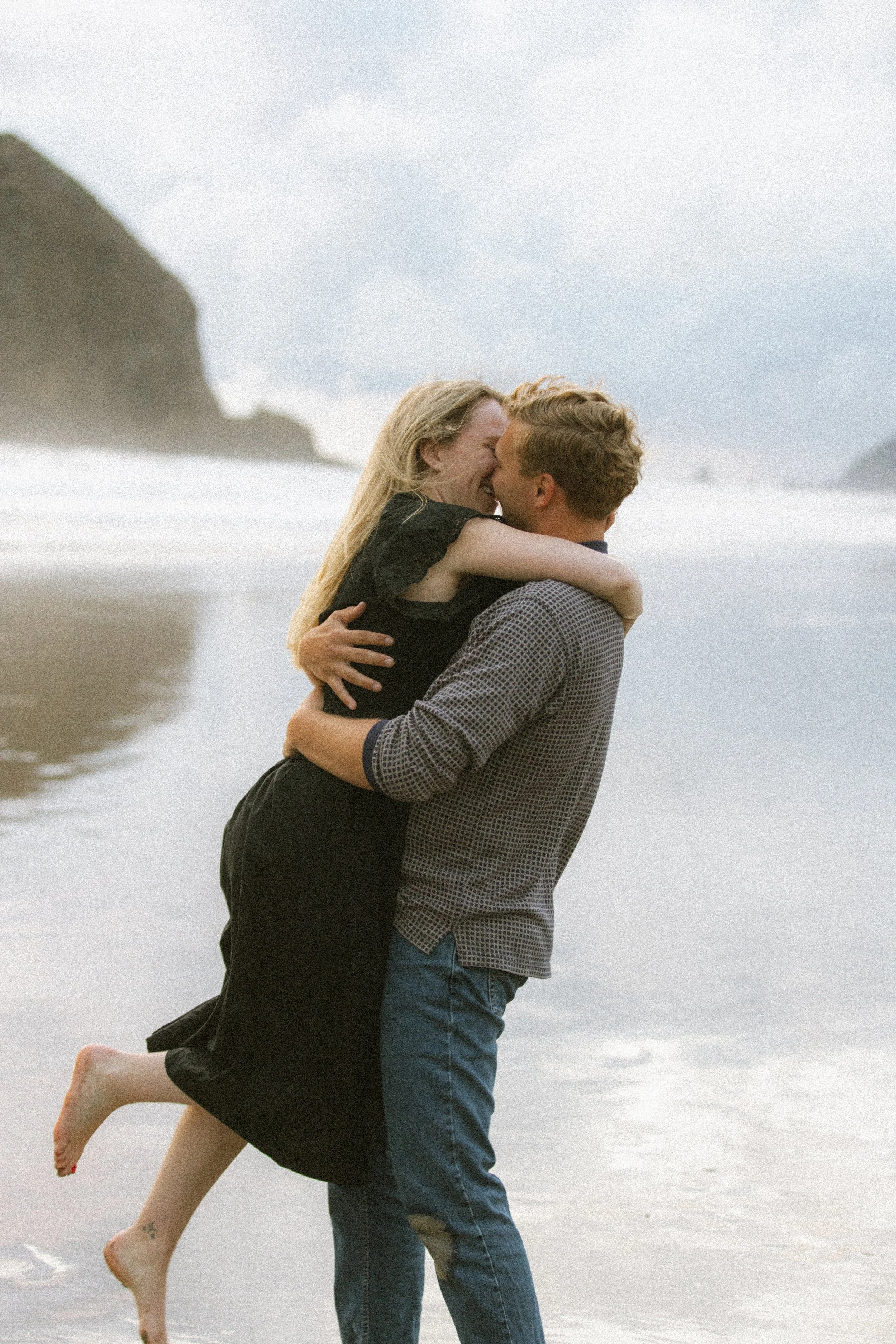 A couple embracing and smiling on the beach, with waves and cliffs in the background.