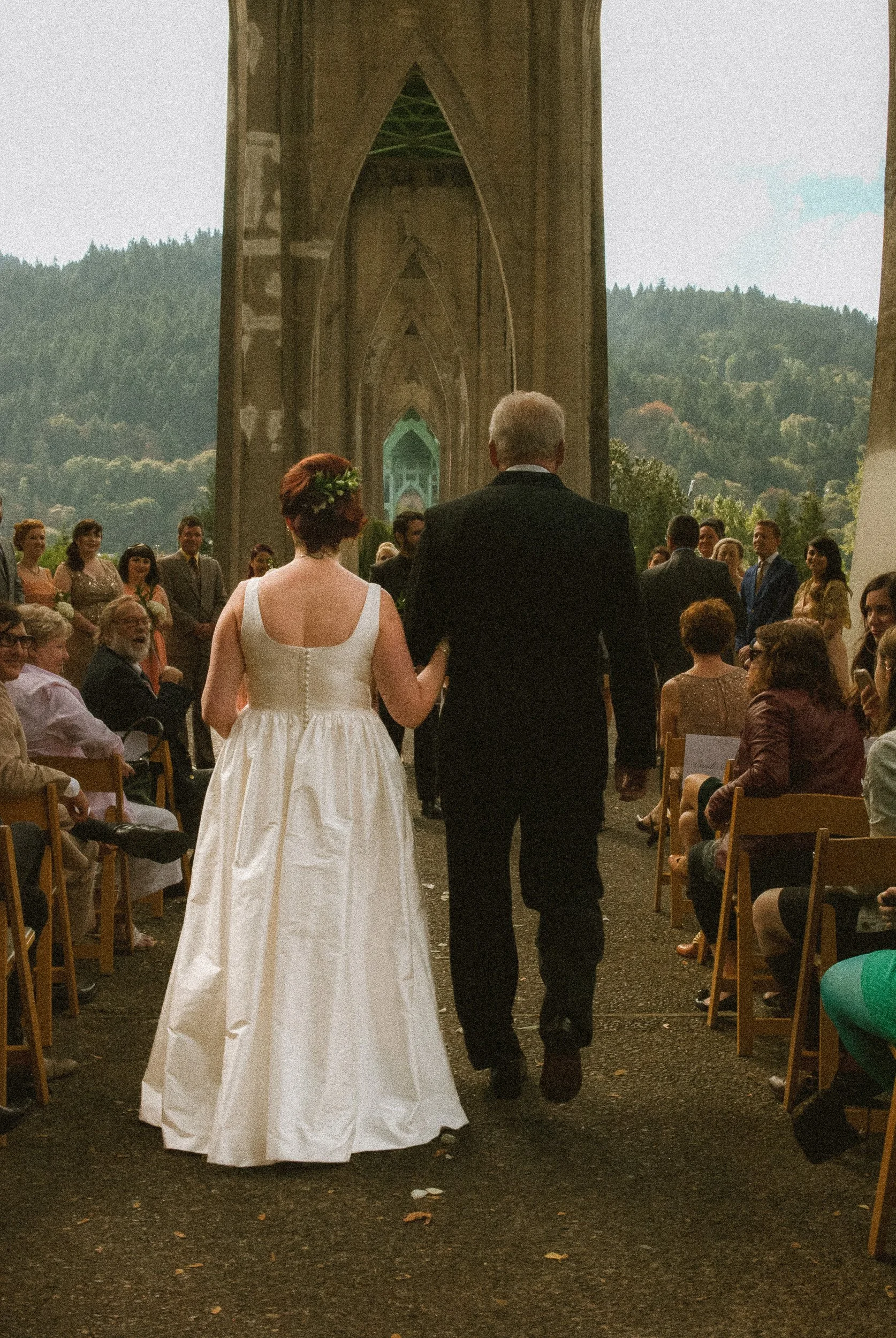 A bride and an older man, likely her father, walking down the aisle of an outdoor wedding ceremony with a Gothic-style stone arch and mountains in the background. The bride is wearing a white wedding gown with a floral crown, and the older man is in 
