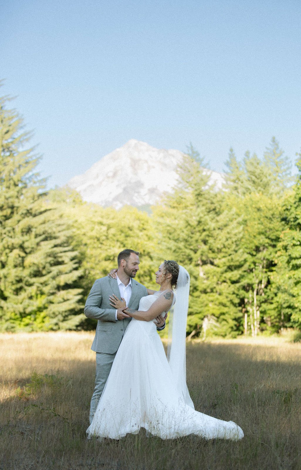 A bride and groom dancing outdoors in a grassy field with trees and snow-capped mountains in the background.