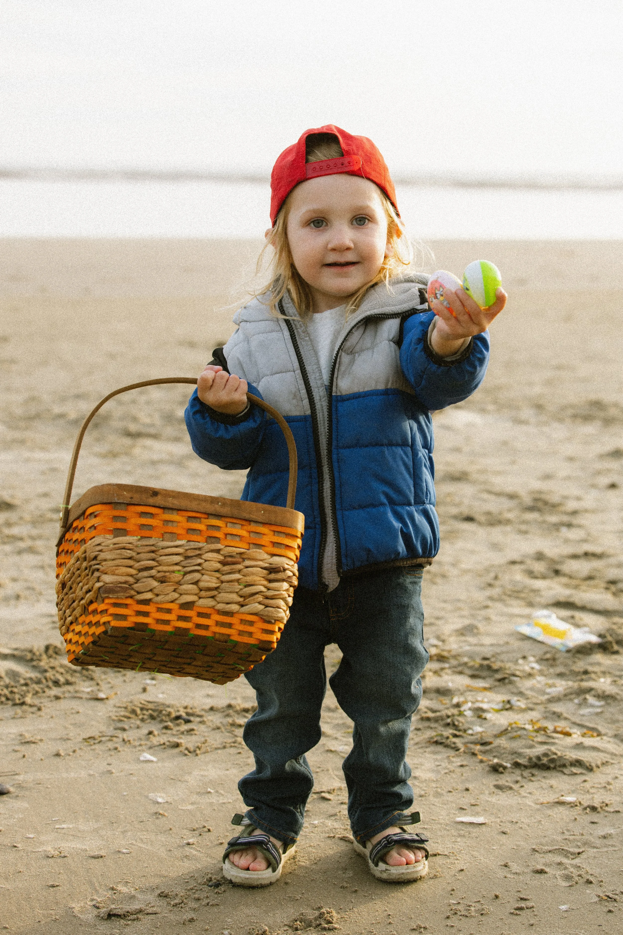 A young boy with blonde hair, wearing a red baseball cap backwards, a blue and gray jacket, and sandals, standing on a beach holding a colorful Easter egg in one hand and a basket in the other.