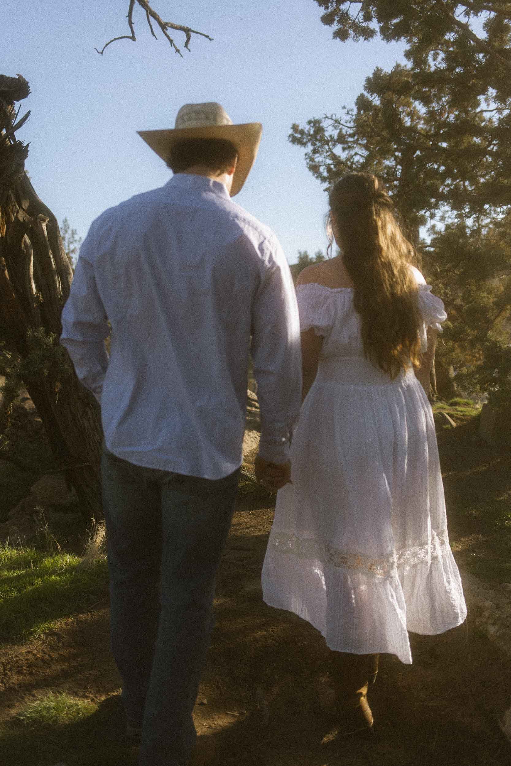 A couple walking hand in hand outdoors, dressed in casual summer clothes, with trees and clear sky in the background.