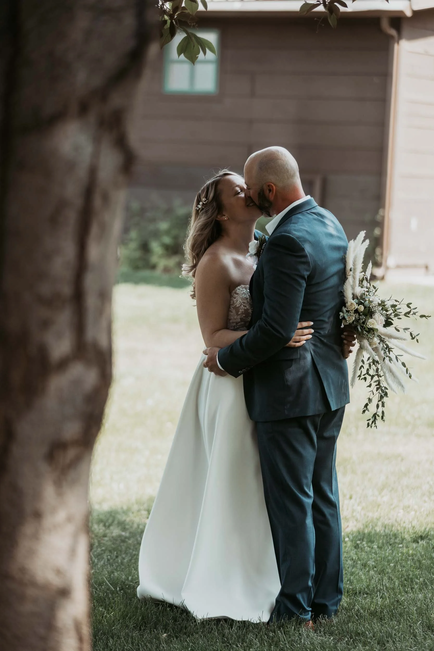 A bride and groom kiss outdoors during their wedding, seen through a tree trunk in the foreground. The bride wears a strapless white wedding gown and the groom wears a dark suit. The groom holds a bouquet of flowers behind his back.