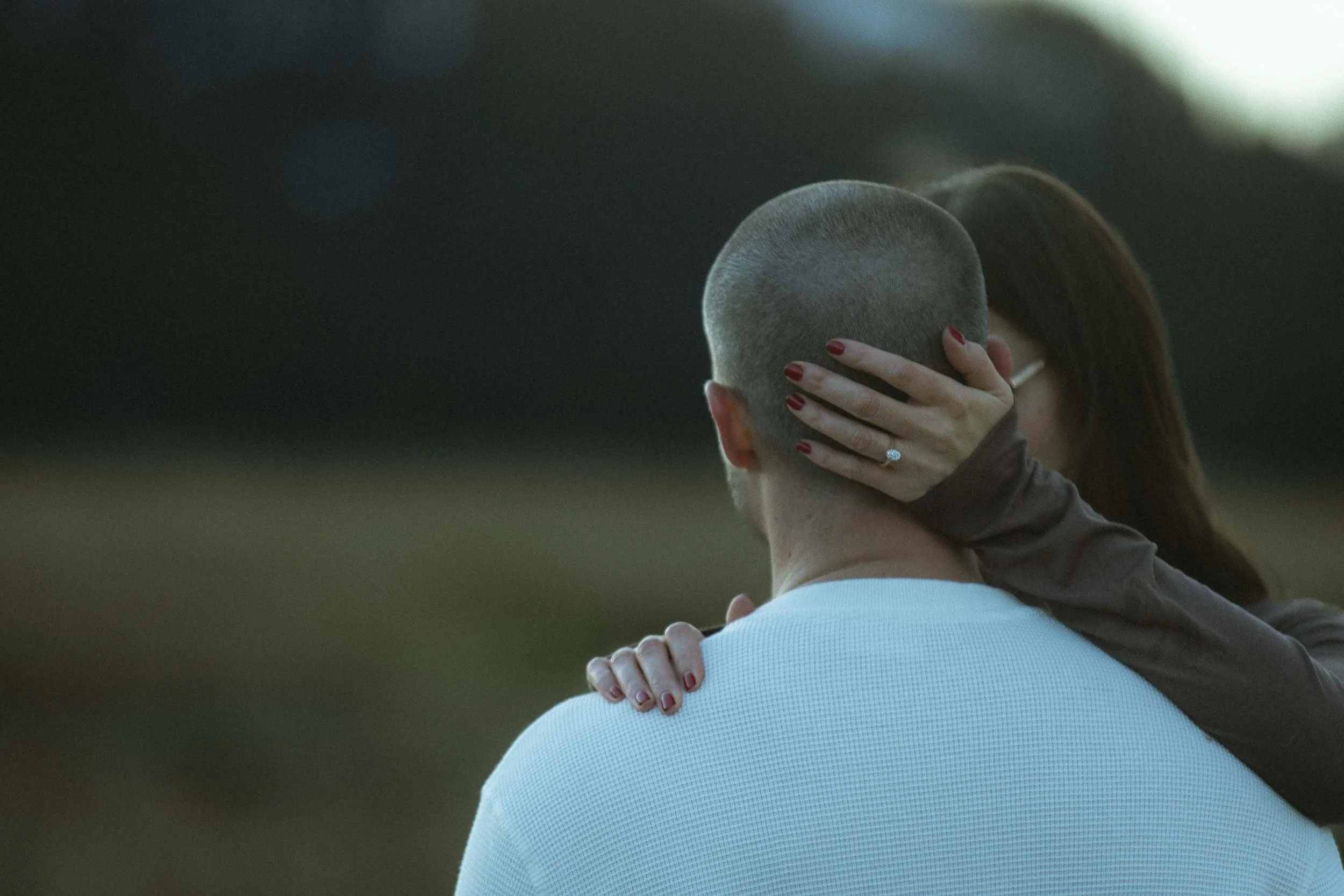 A woman with red painted nails and wearing glasses gently touches the shaved head of a man wearing a white shirt outdoors.