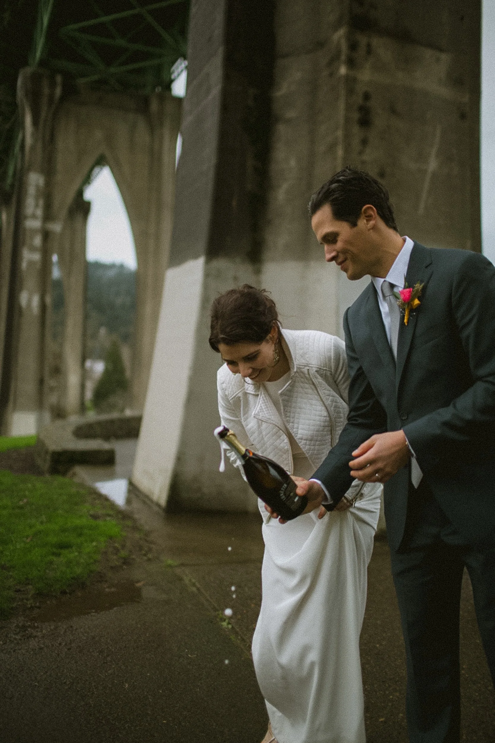 A couple celebrating outdoors under a bridge, with the woman holding a champagne bottle and both smiling.