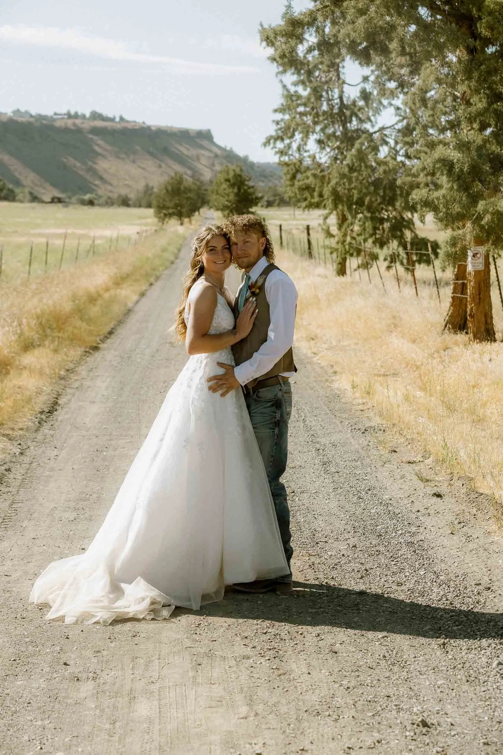 A bride and groom stand in a grassy outdoor setting with water and trees in the background, illuminated by sunlight, during their wedding ceremony.