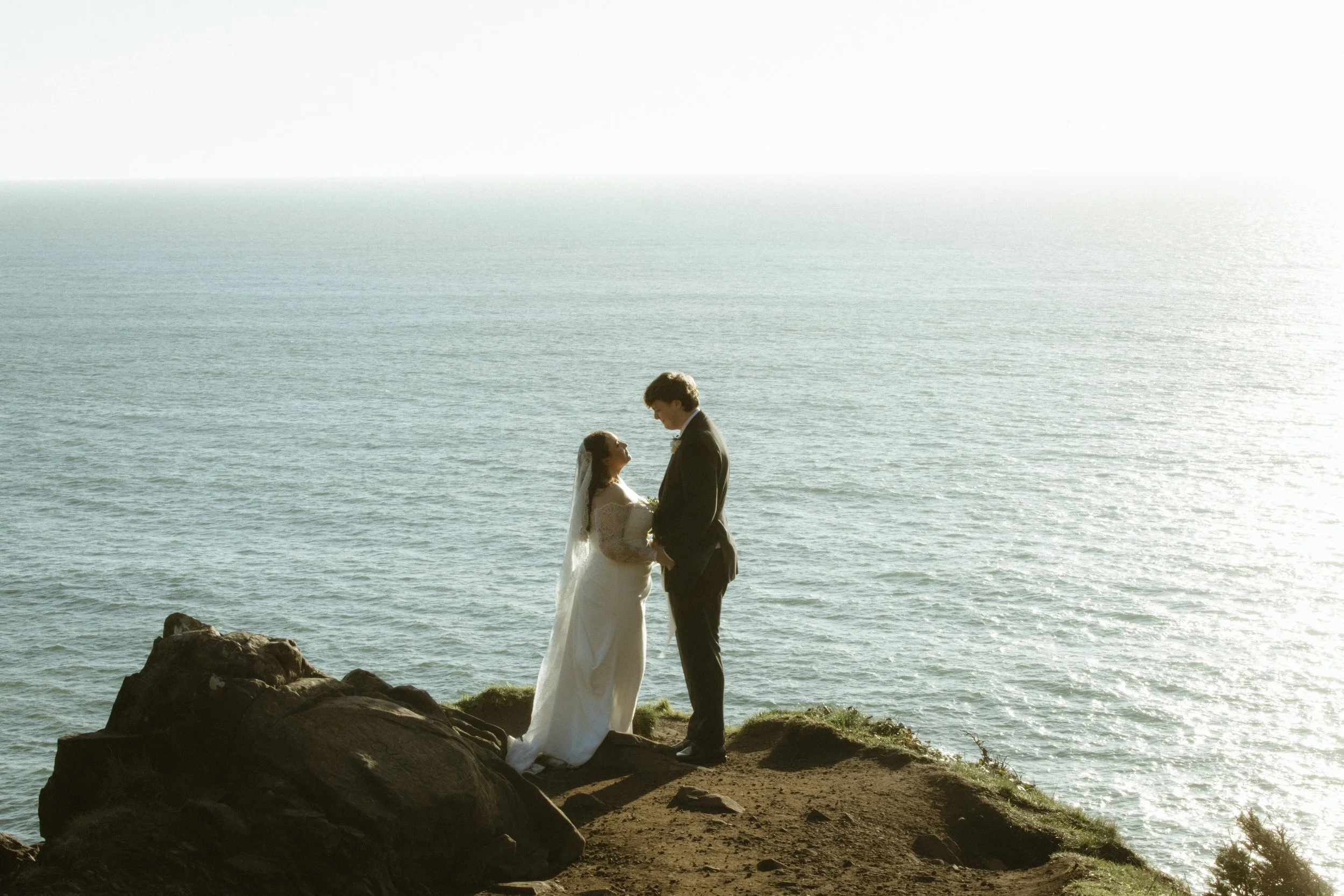A bride and groom holding hands and facing each other on a rocky beach by the ocean during sunset or sunrise.