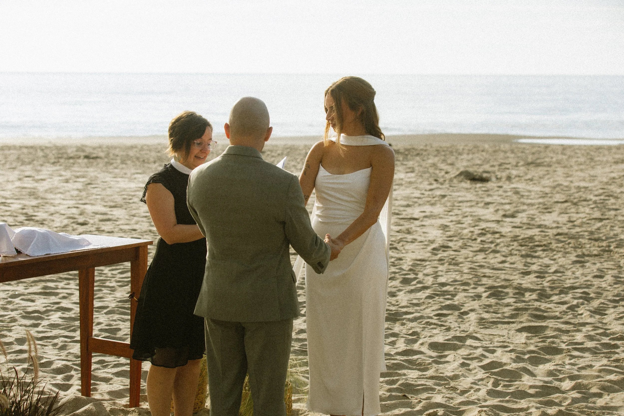 A beach wedding ceremony with a bride and groom holding hands in front of an officiant and celebrating the moment.