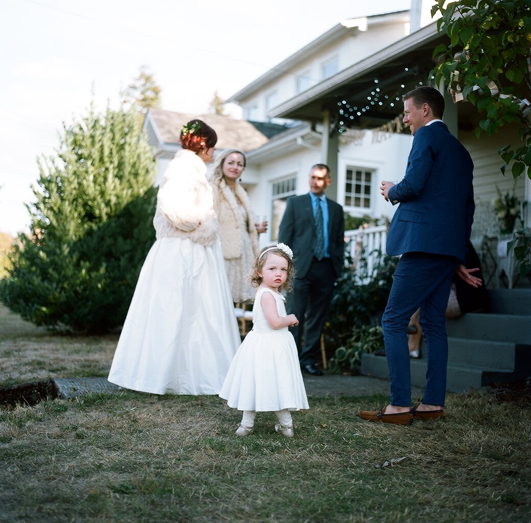 People gathered outside a house for a wedding celebration. A young girl in a white dress stands in the foreground, while a woman in a white gown and a man in a suit are in conversation with a man in a blue suit. Part of a house with string lights han