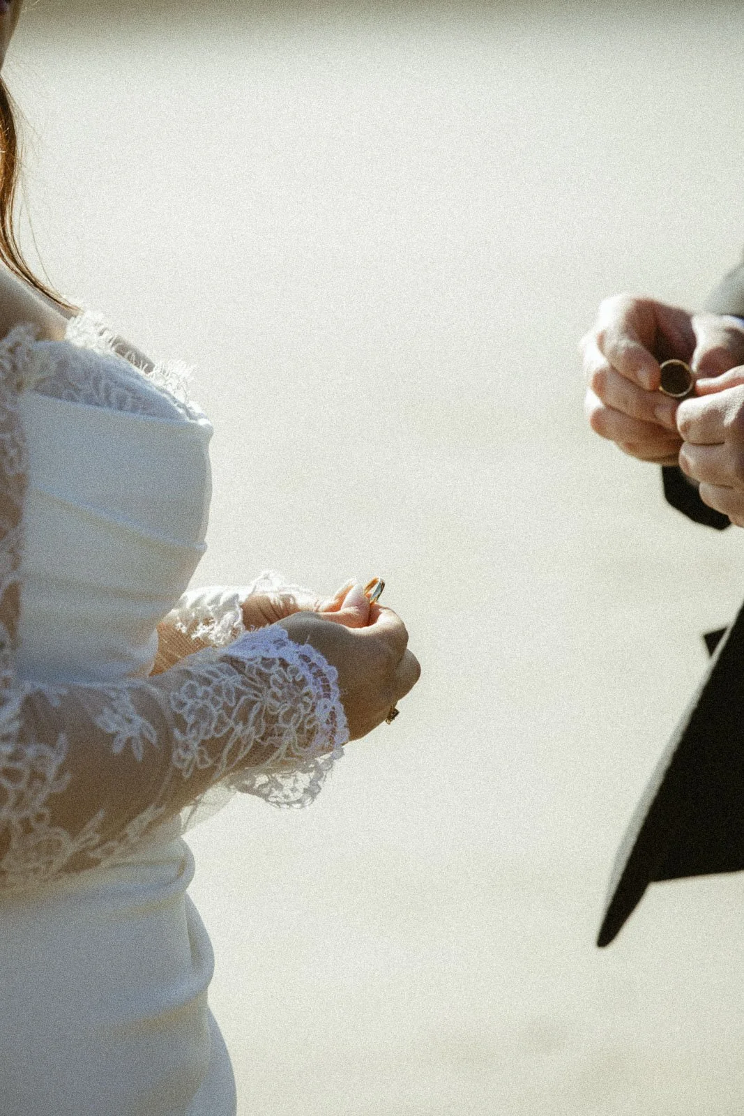 Close-up of a wedding ceremony with a bride in a lace dress and a groom in a suit exchanging rings outside.