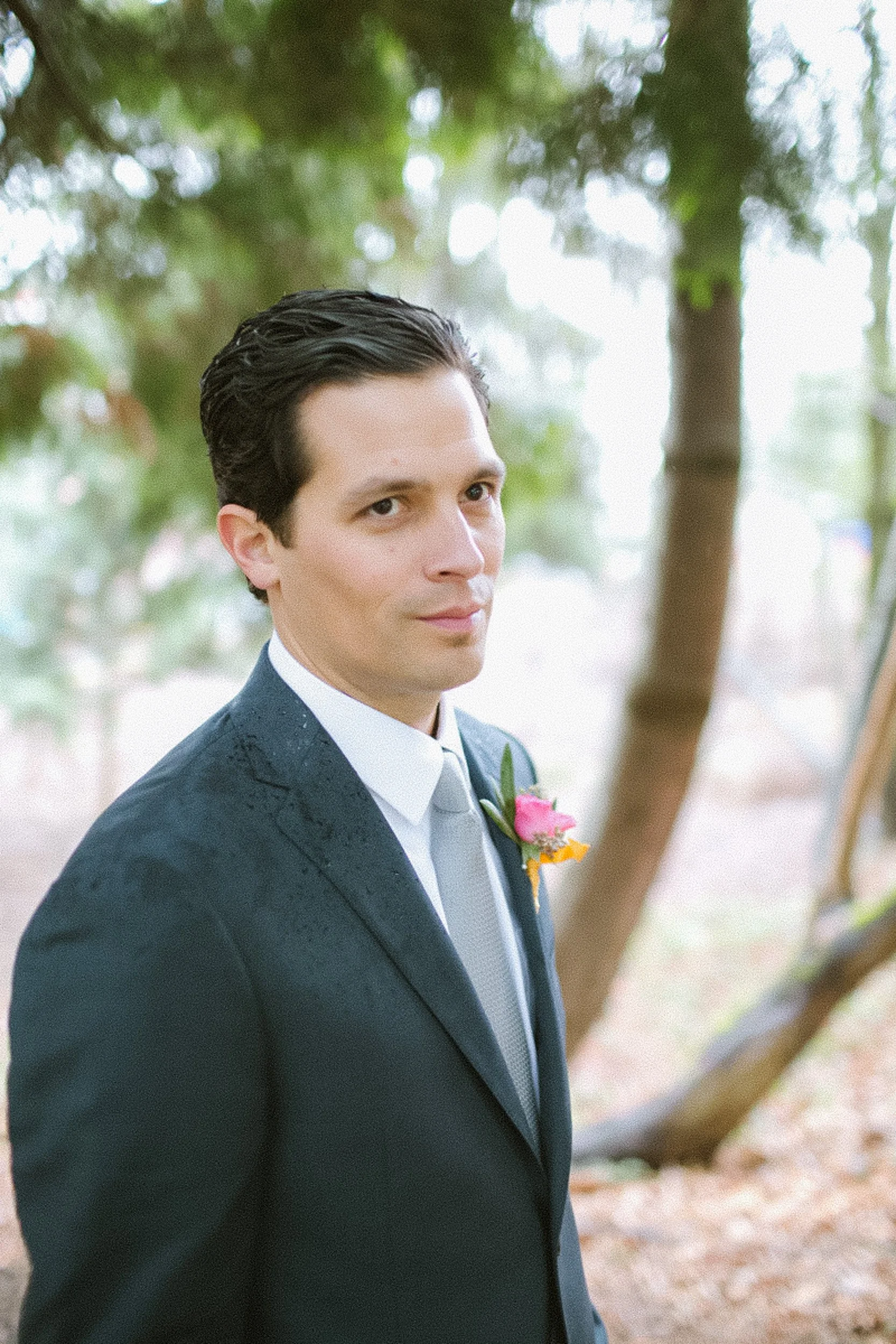 A man in a black suit with a white shirt and tie, standing outdoors near trees, with a pink and yellow boutonniere on his lapel.