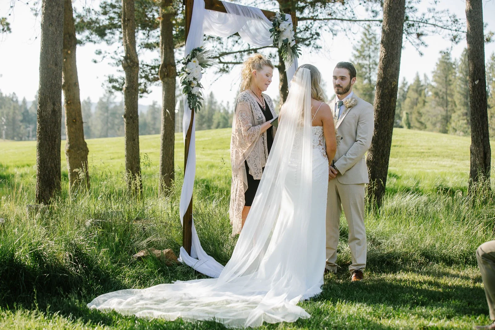 A wedding ceremony taking place outdoors in a forested area with grass and tall trees. The bride and groom stand in front of the officiant under a decorated archway with flowers and white fabric. The bride wears a lace wedding dress with a long veil, and the groom wears a light-colored suit. The officiant reads from a book as the couple exchanges vows.