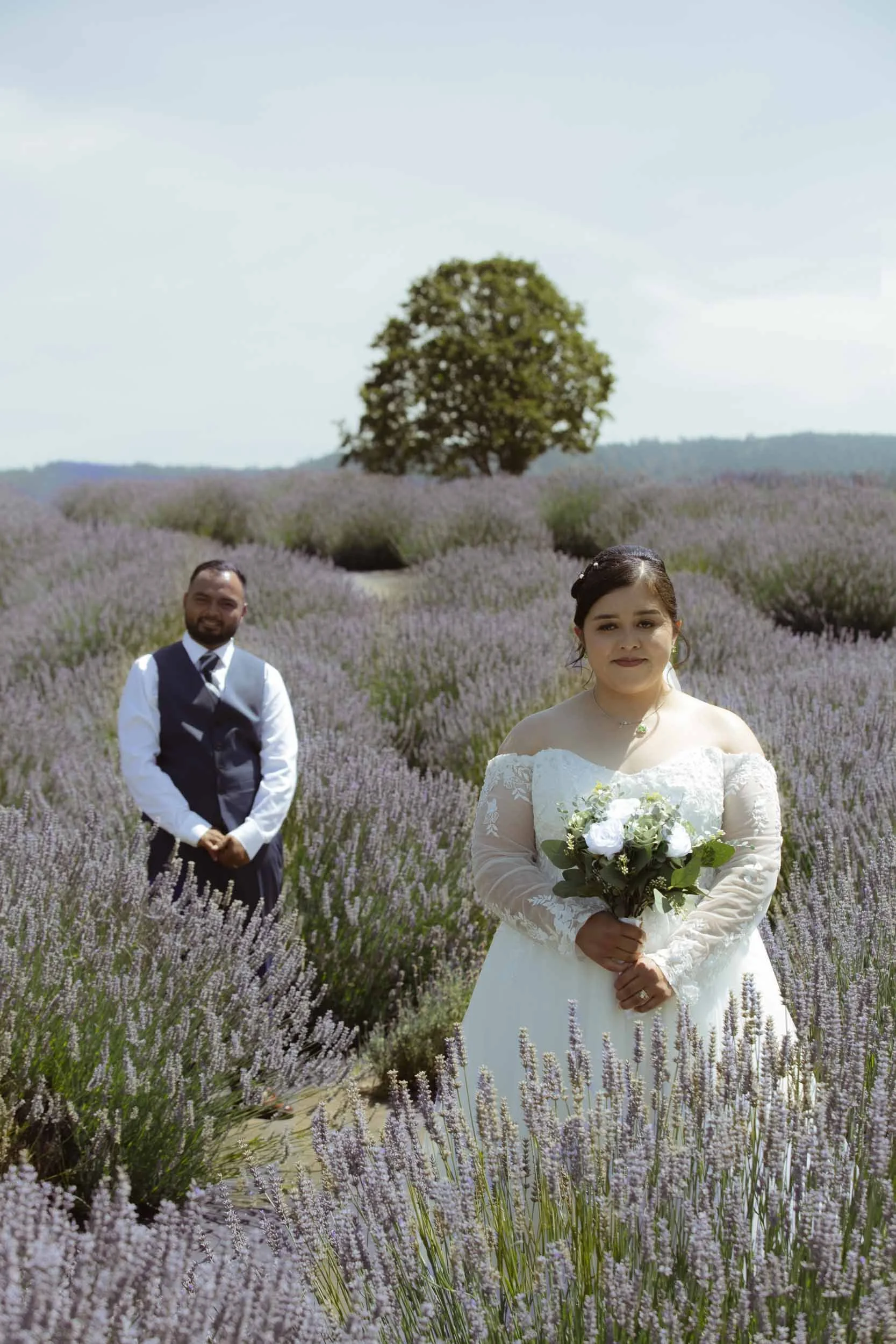 A bride holding a bouquet of white roses in a lavender field with a groom in the background, and a large tree in the distance under a partly cloudy sky.