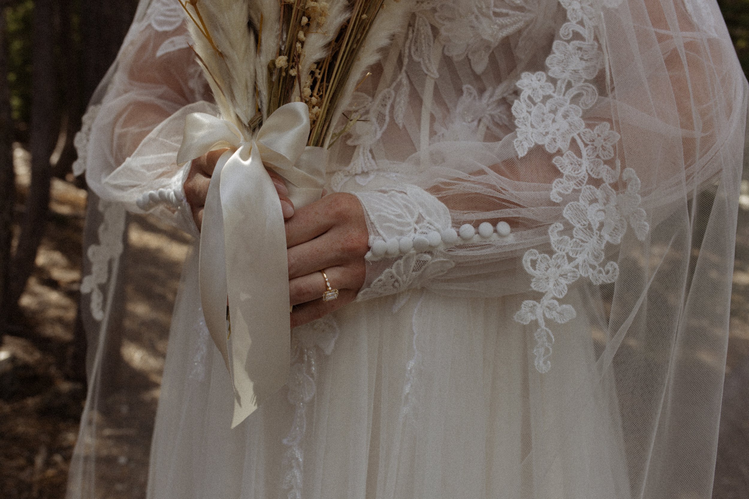 Close-up of a person in a lace wedding dress holding a bouquet of dried flowers tied with a satin ribbon, with a ring on their finger.