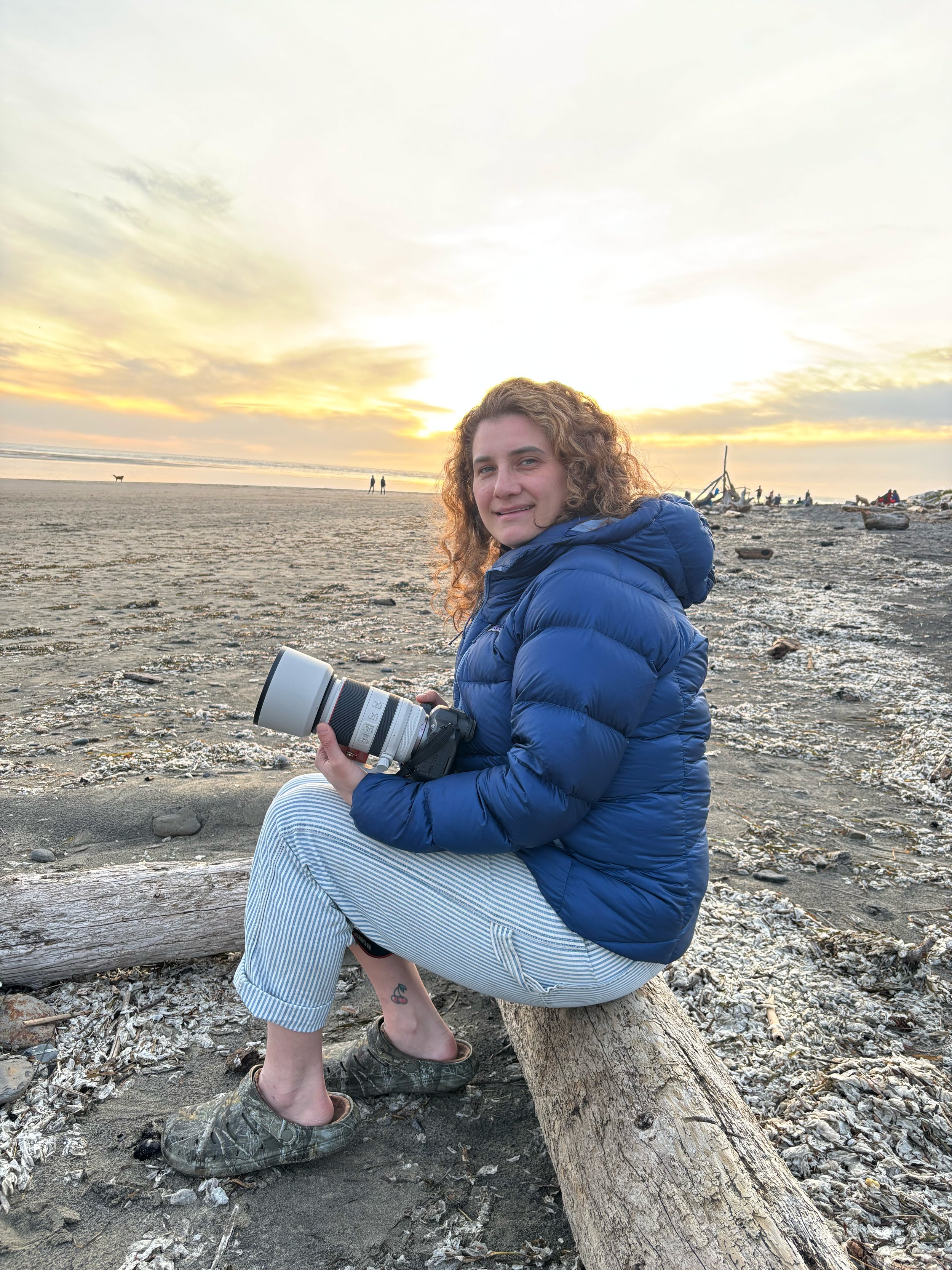A woman with curly hair sitting on a log on the beach during sunset, holding a camera, wearing a blue puffer jacket, striped pants, and camo shoes, with a small tattoo of cherries on her ankle.
