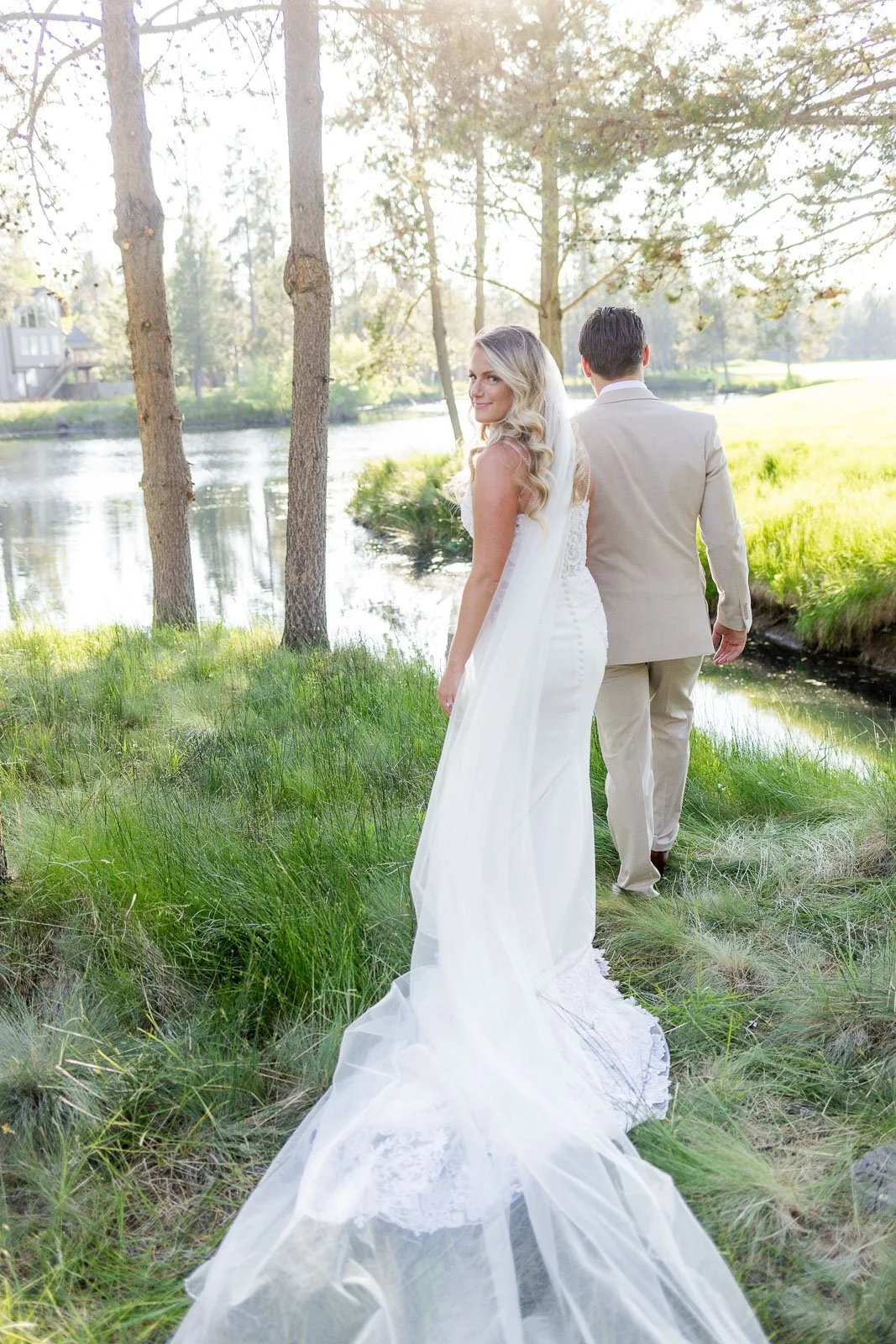 A bride in a white wedding dress with a long train, looking back over her shoulder, standing near a river with trees and a grassy area, with a groom in a light-colored suit walking ahead of her.