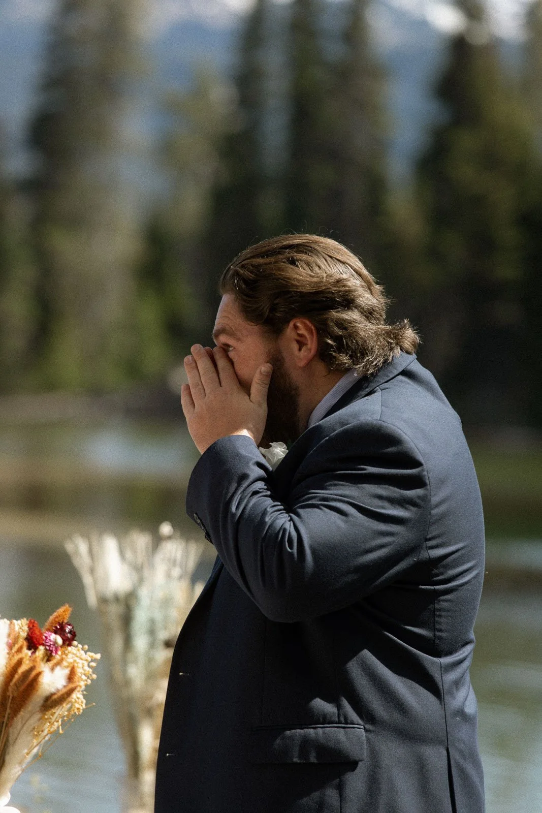 A man with long hair and a beard, dressed in a navy suit, is standing outdoors with his hands covering his face, appearing emotional. Background is blurred with trees and water.