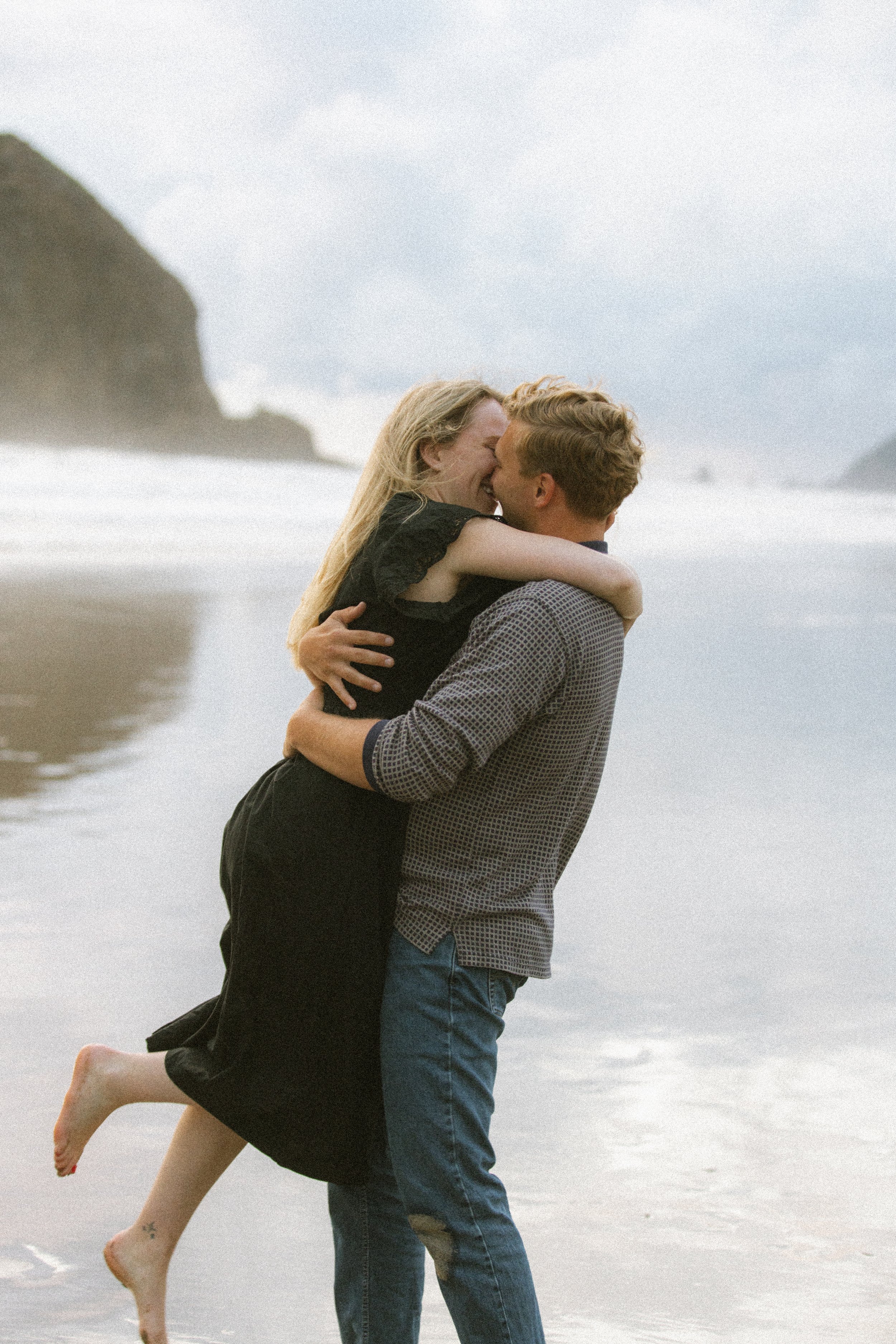A couple embracing and laughing on the beach with cliffs in the background.