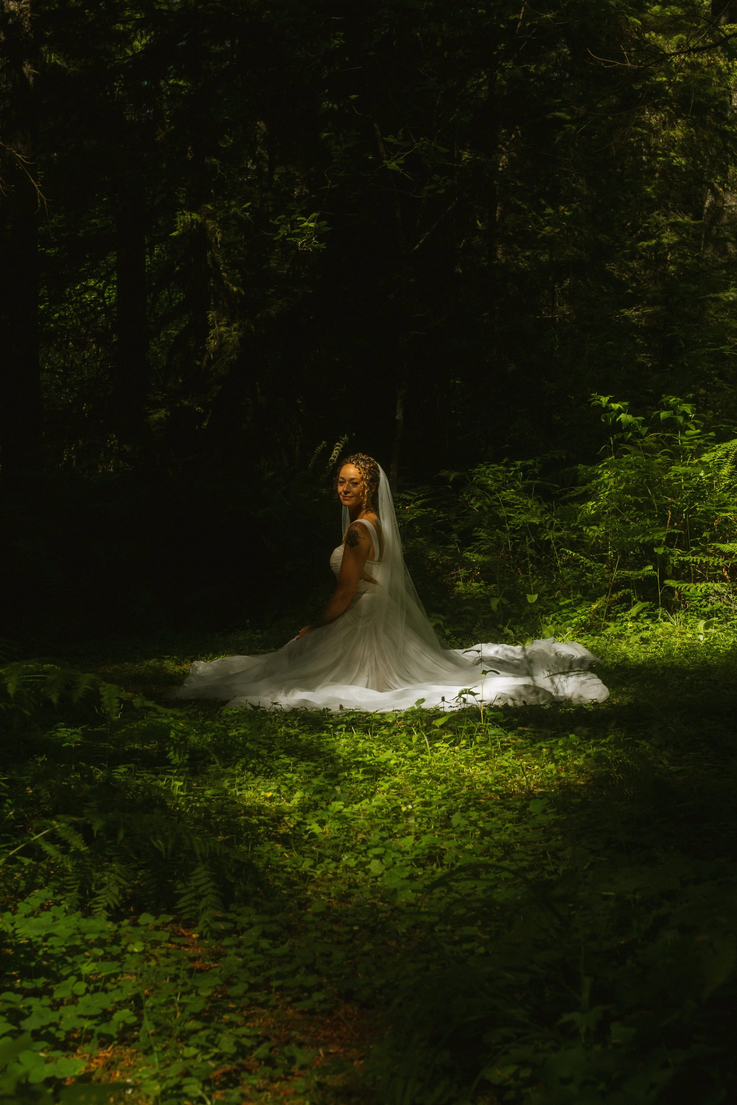 A woman in a white wedding dress and veil sitting on the ground in a forest surrounded by green foliage, with sunlight filtering through the trees.