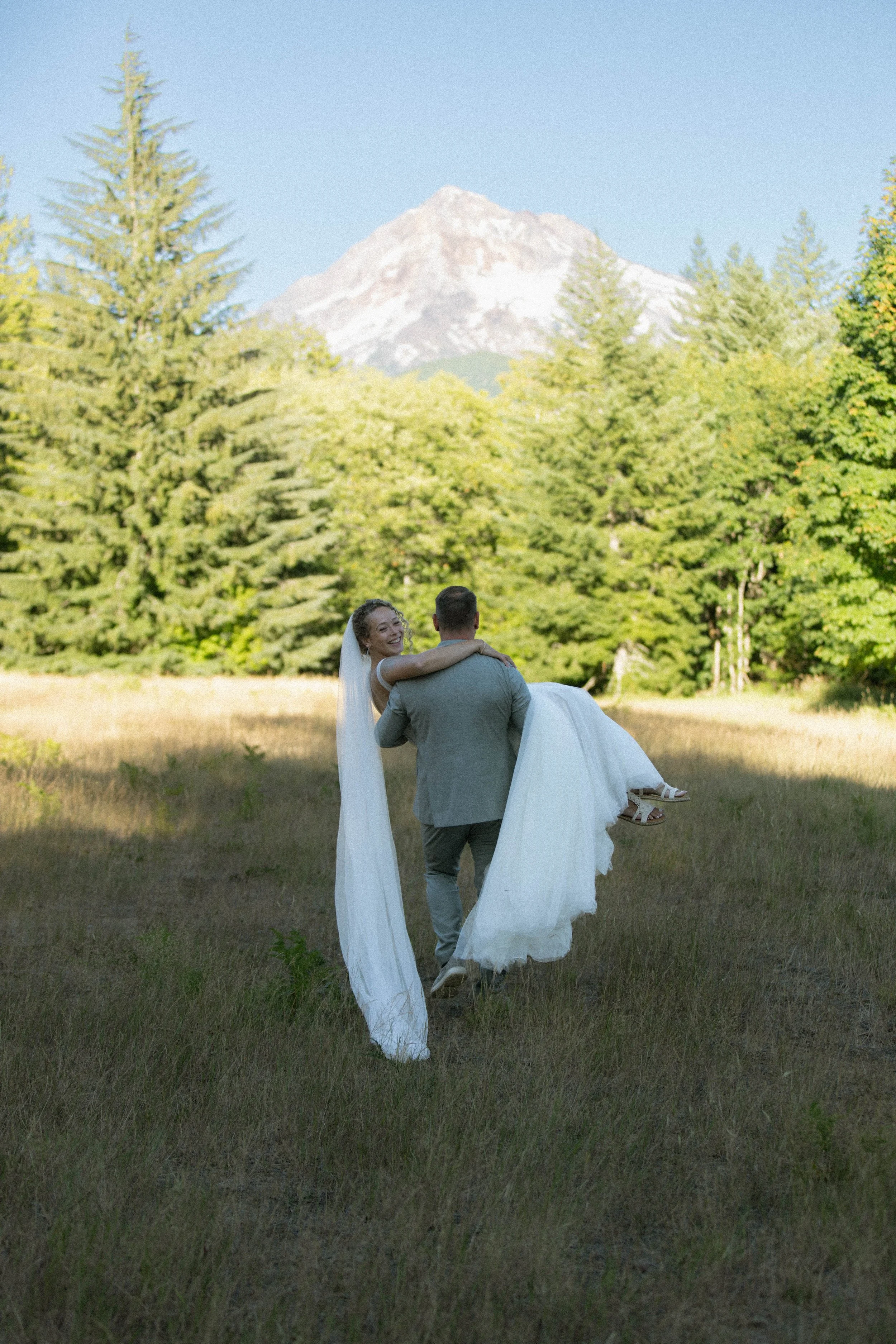 A bride being carried by a groom in a scenic outdoor setting with a mountain and trees in the background.